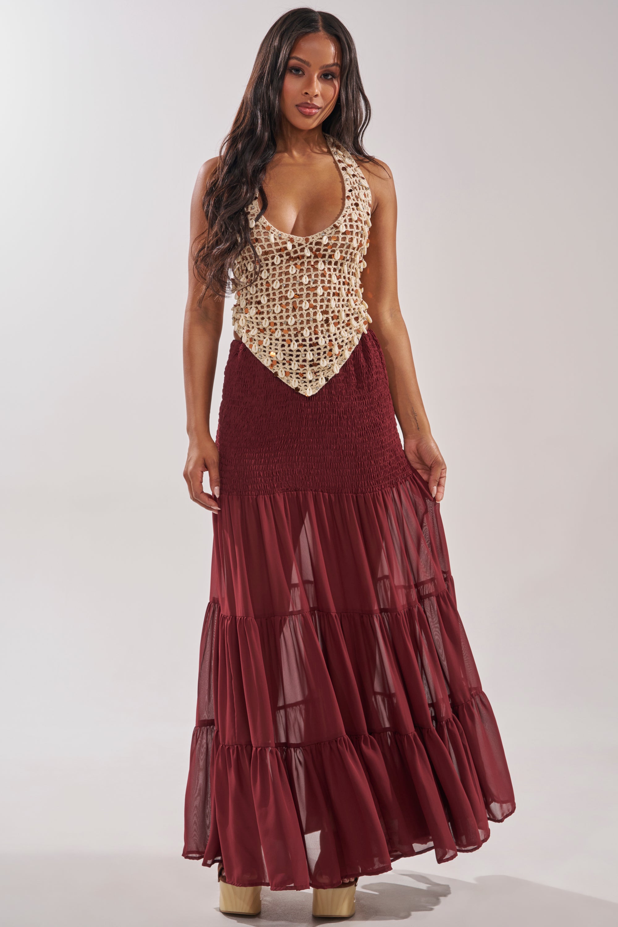A woman with long, wavy hair wears platform shoes and a sleeveless beige crochet top with a deep neckline. She stands facing forward in the DRIFTING AWAY MAXI SKIRT, a flowy, tiered maroon piece, against a plain light gray background. 5