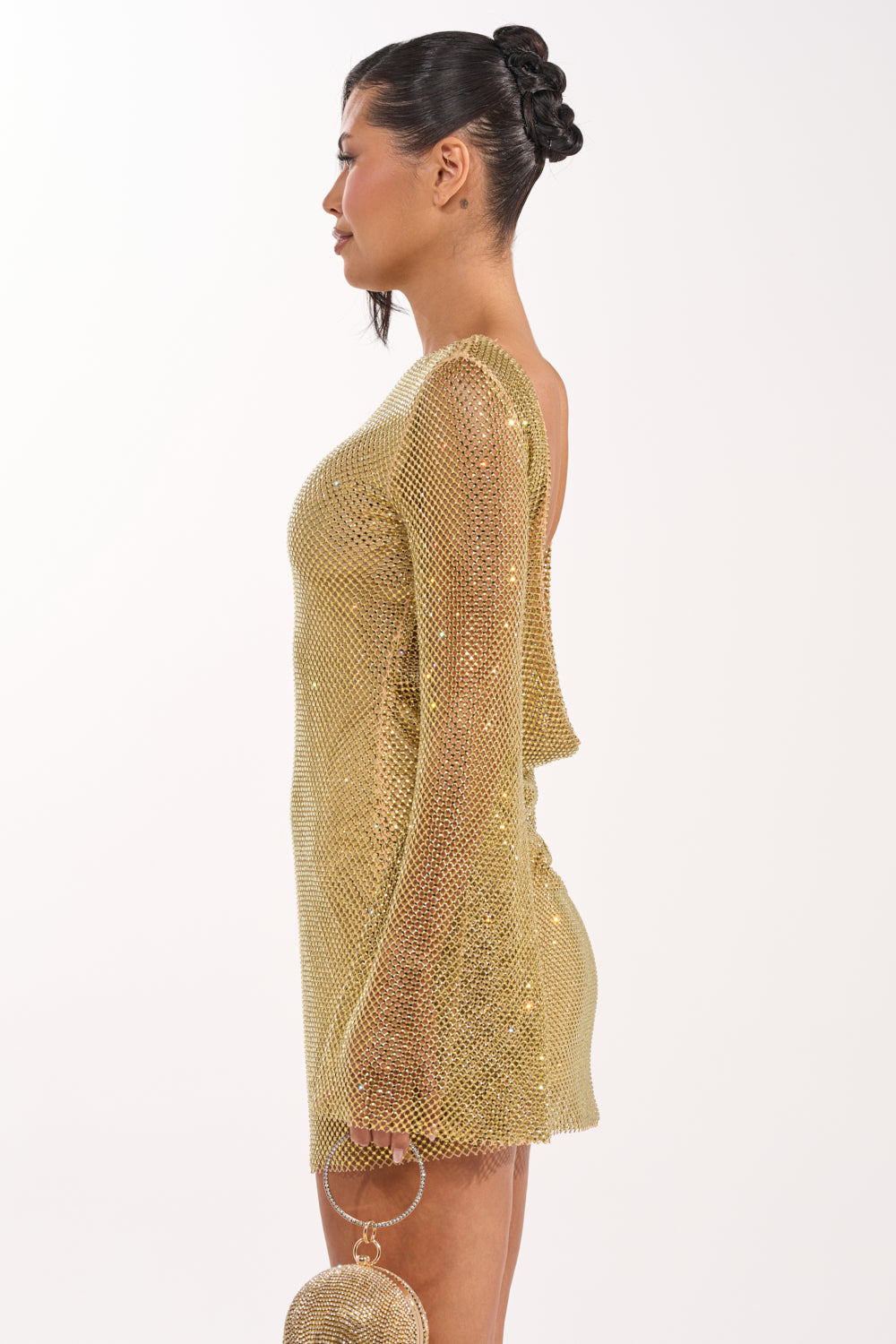 Woman in profile view wearing the TAKE ME TO IBIZA RHINESTONE MINI DRESS, a gold mesh mini with long sleeves and open back, holding a round gold handbag. Her dark hair is styled elegantly against a plain white background. 7
