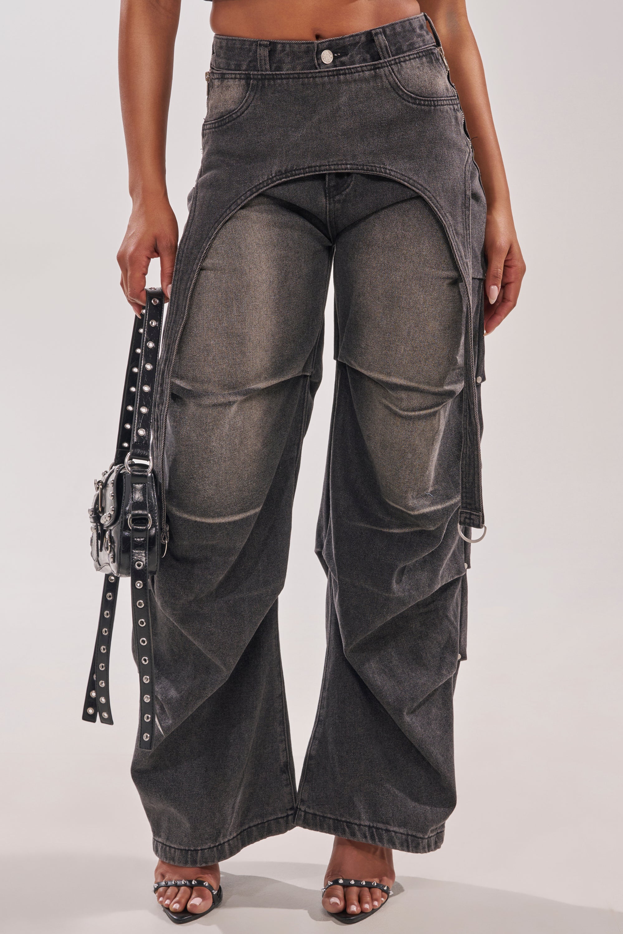 A person in utility-inspired streetwear: dark-wash LEGEND TROUSER with unique hip overlay, holding a black studded handbag and wearing heeled sandals; photo cropped at the chest. 2
