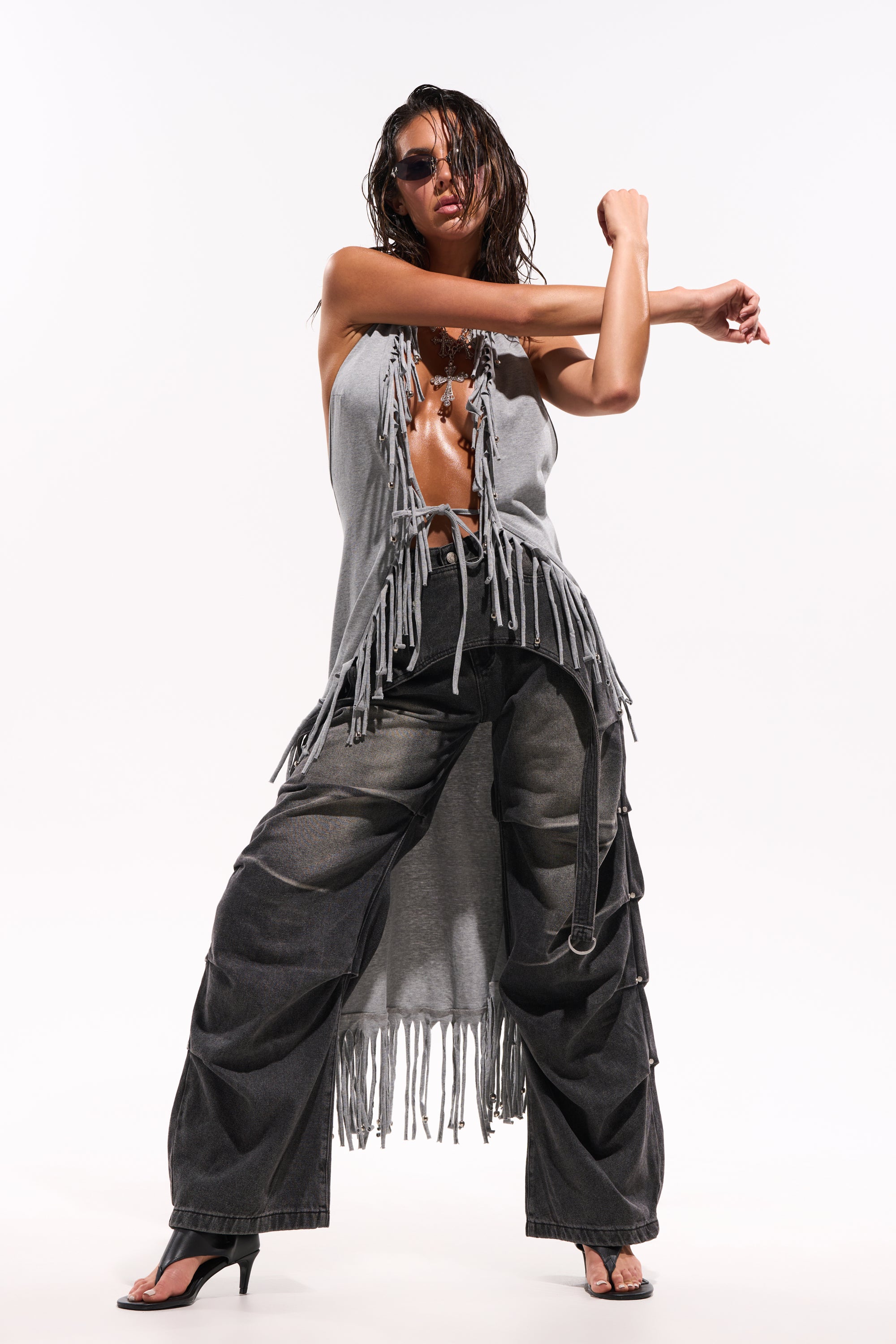A person in the LEGEND TROUSER, black wide-leg cargo denim pants, and a sleeveless gray fringed top poses confidently in heeled sandals and sunglasses against a white background, with one arm stretched across their chest. 1