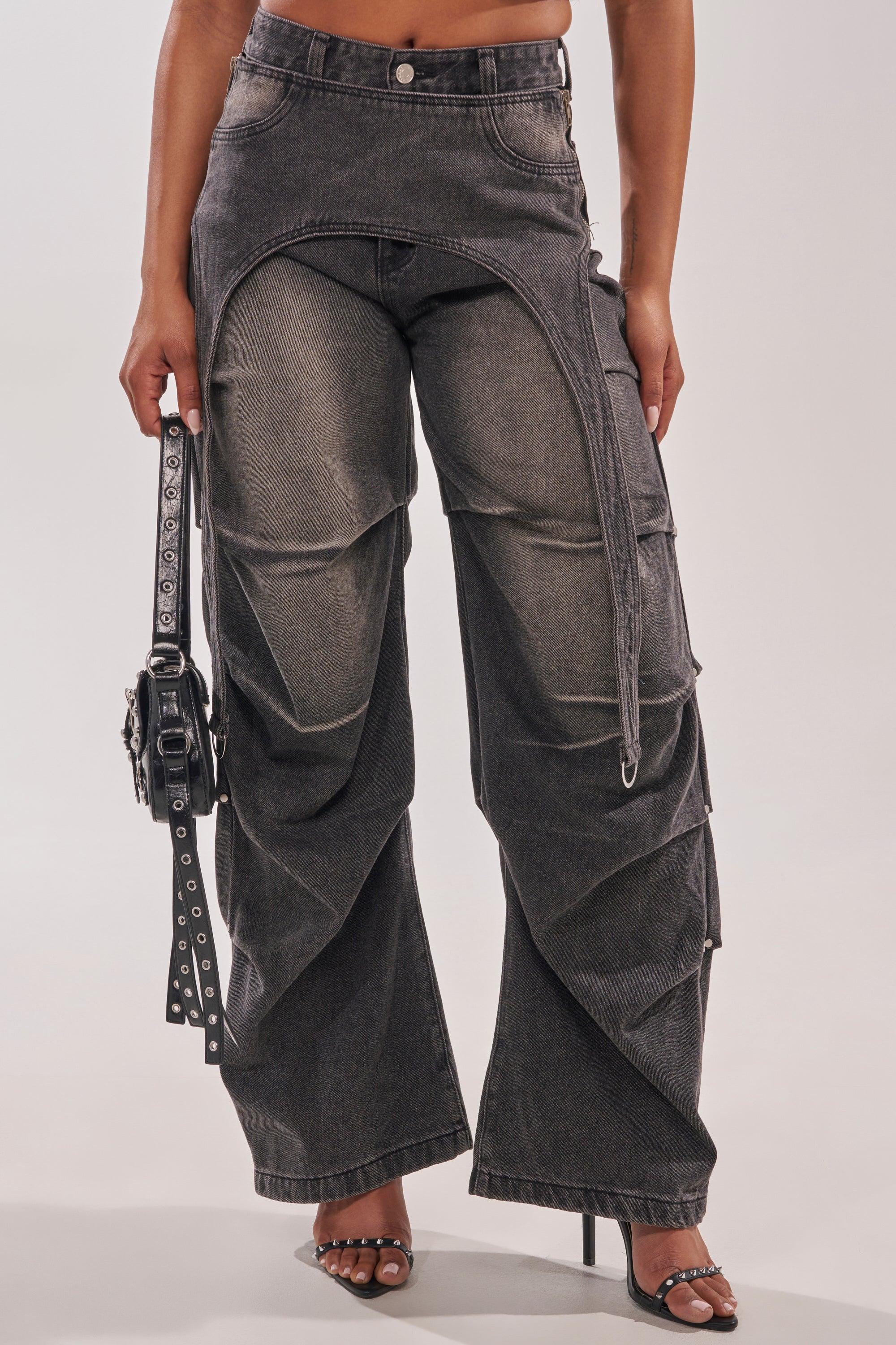 Wearing the LEGEND TROUSER—dark gray denim with a unique flap detail—paired with a black studded handbag and high-heeled sandals, the outfit creates a bold western streetwear look. 4