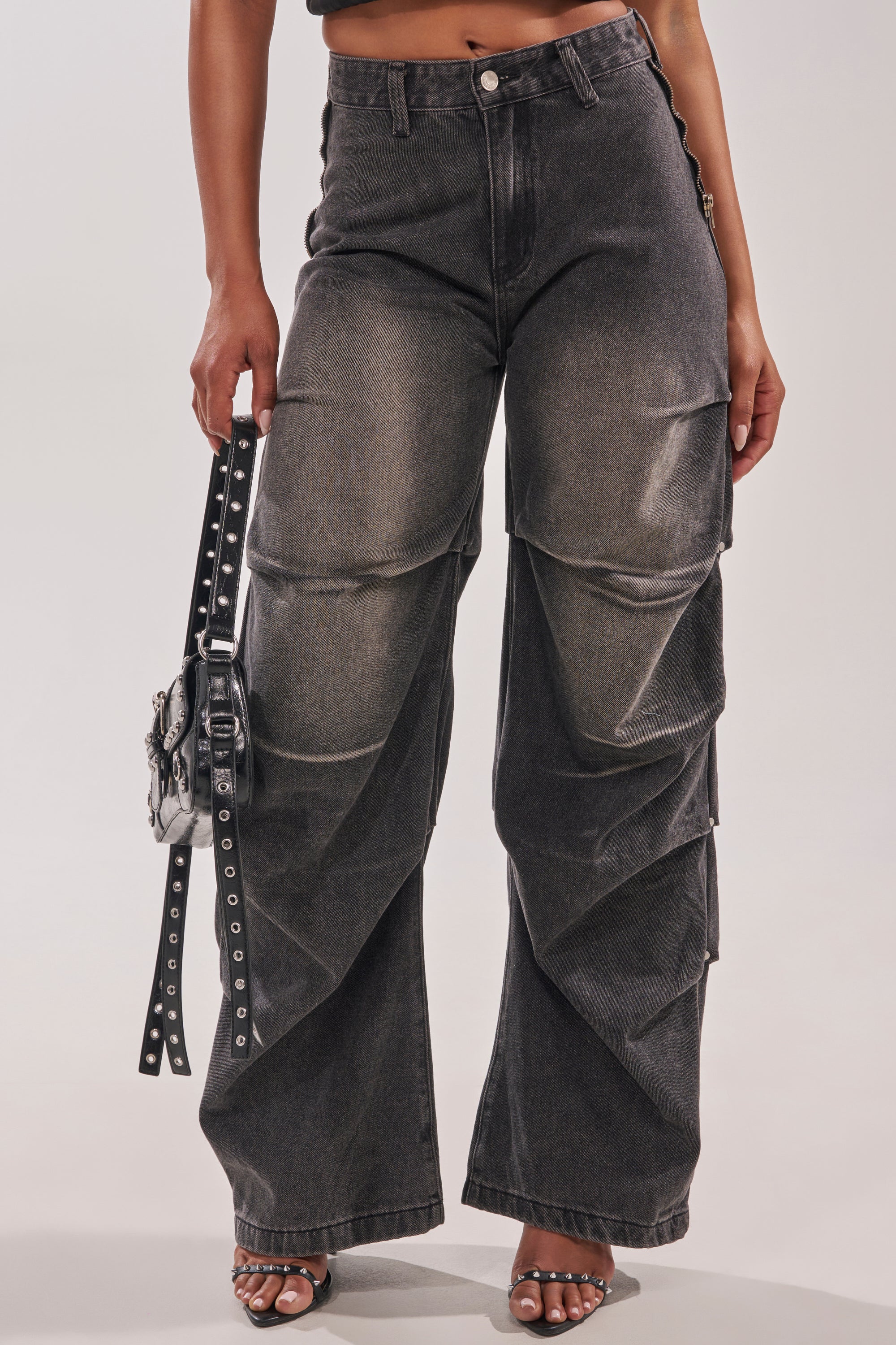 Wearing the LEGEND TROUSER—high-waisted, wide-leg, dark wash jeans with layered western streetwear details—paired with a small black studded handbag and heeled sandals against a plain light background. 5