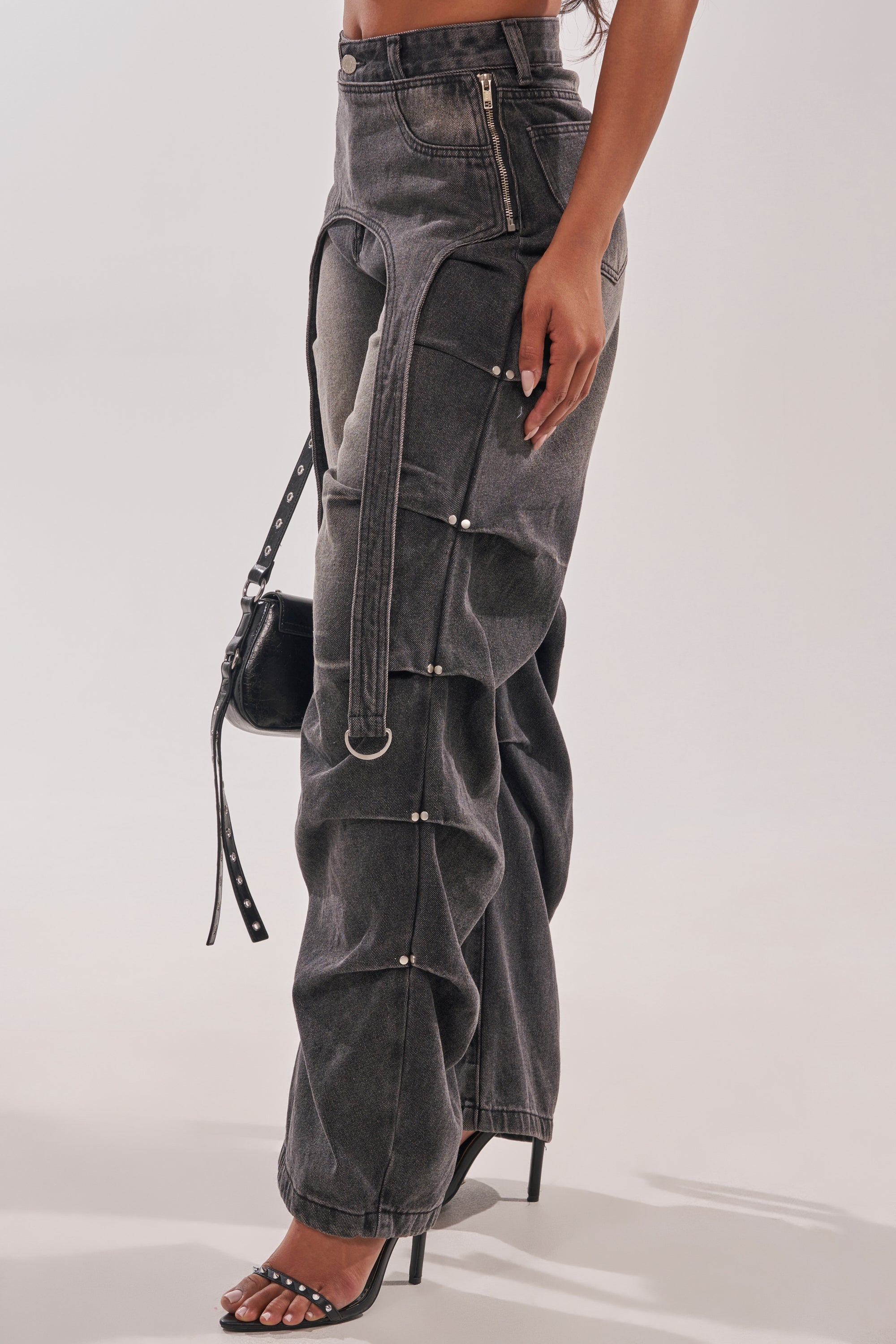 A person styled in LEGEND TROUSER—a black wide-leg cargo denim pant with strap and button details—paired with heeled sandals and a small black handbag for a chic, utility-inspired streetwear look. 6