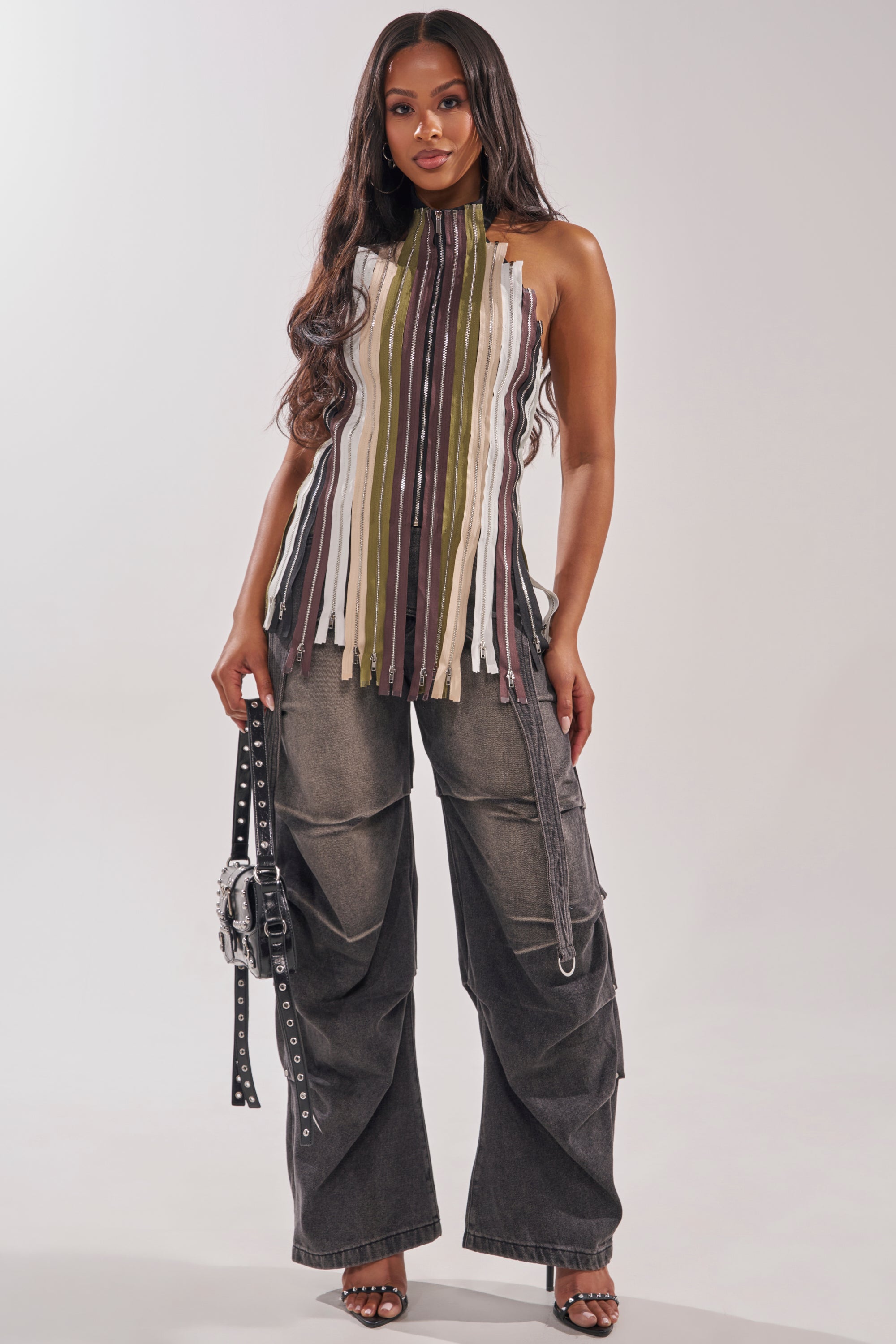 A woman with long wavy hair wears a halter top with vertical multicolored fringe strips and the LEGEND TROUSER, black wide-leg cargo denim pants. She holds a studded bag and strappy heeled sandals against a plain light background. 9