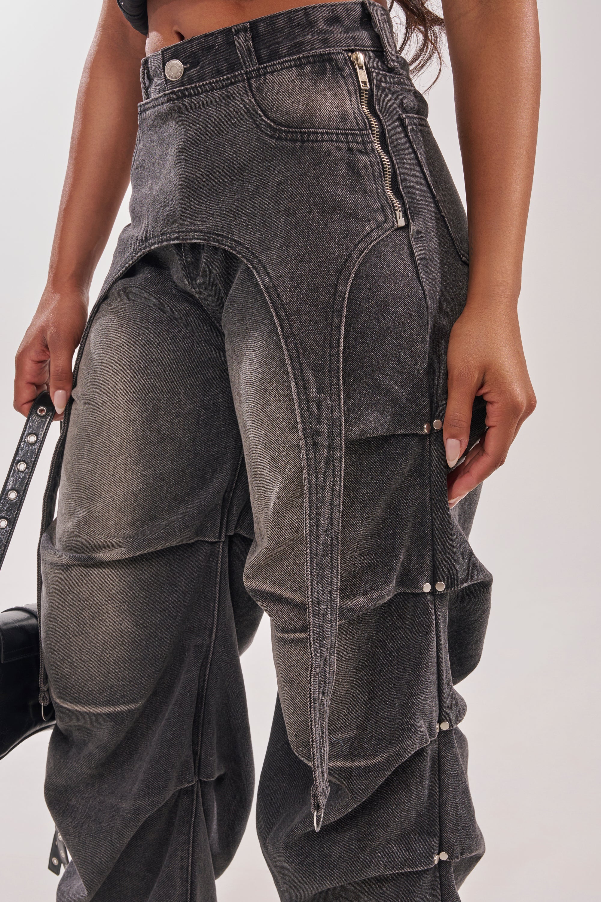 Wearing the LEGEND TROUSER—dark gray, high-waisted, distressed jeans with layered panels, visible zippers, and silver buttons—a person stands against a plain light background. 10