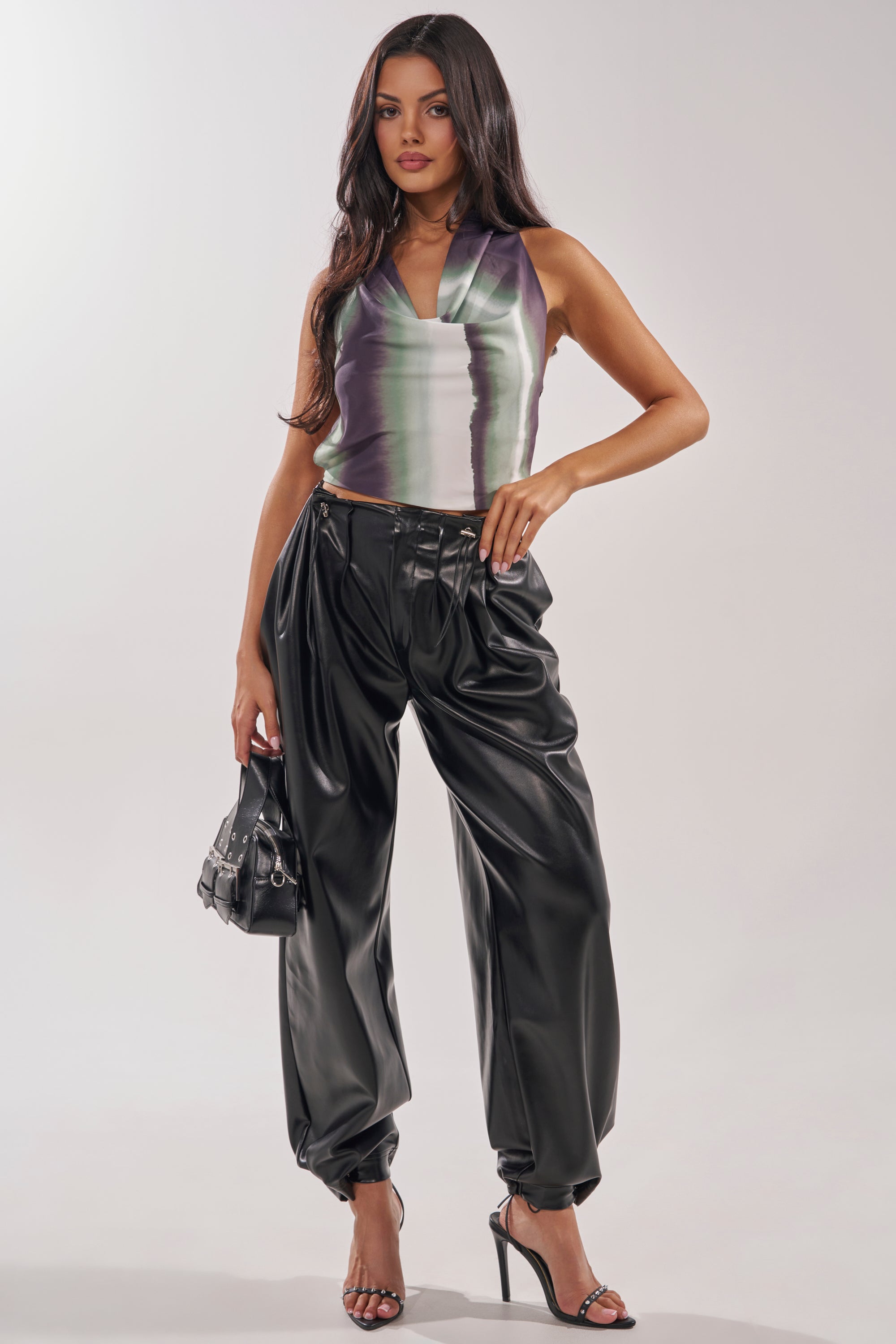 A woman wears a sleeveless satin halter top with a green and purple gradient, paired with the ON TOP OF IT FAUX LEATHER TROUSER in black, holding a small black handbag and wearing black high-heeled sandals. 0