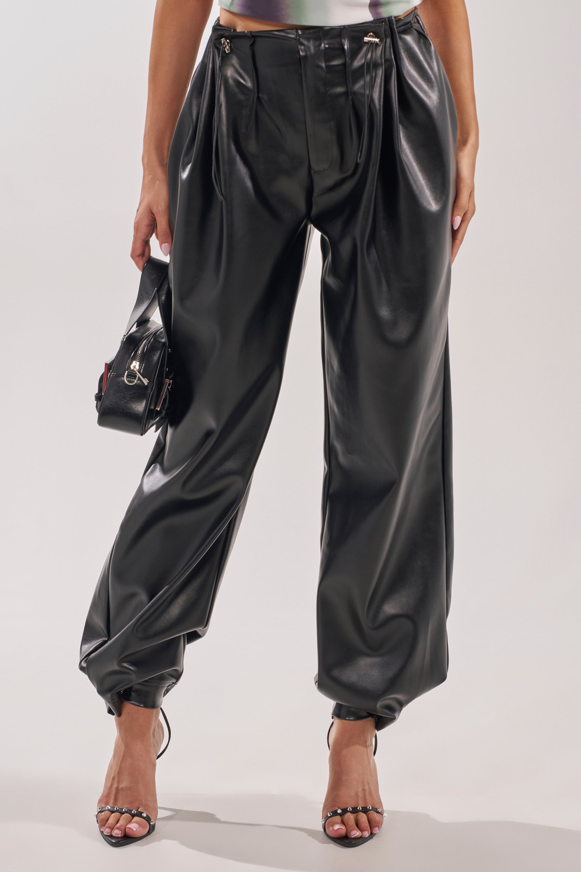 A person wears the ON TOP OF IT FAUX LEATHER TROUSER—high-rise, loose-fitting black wide-leg pants with pleats—paired with strappy black heels and a small black handbag. The image is cropped above the chest. 1