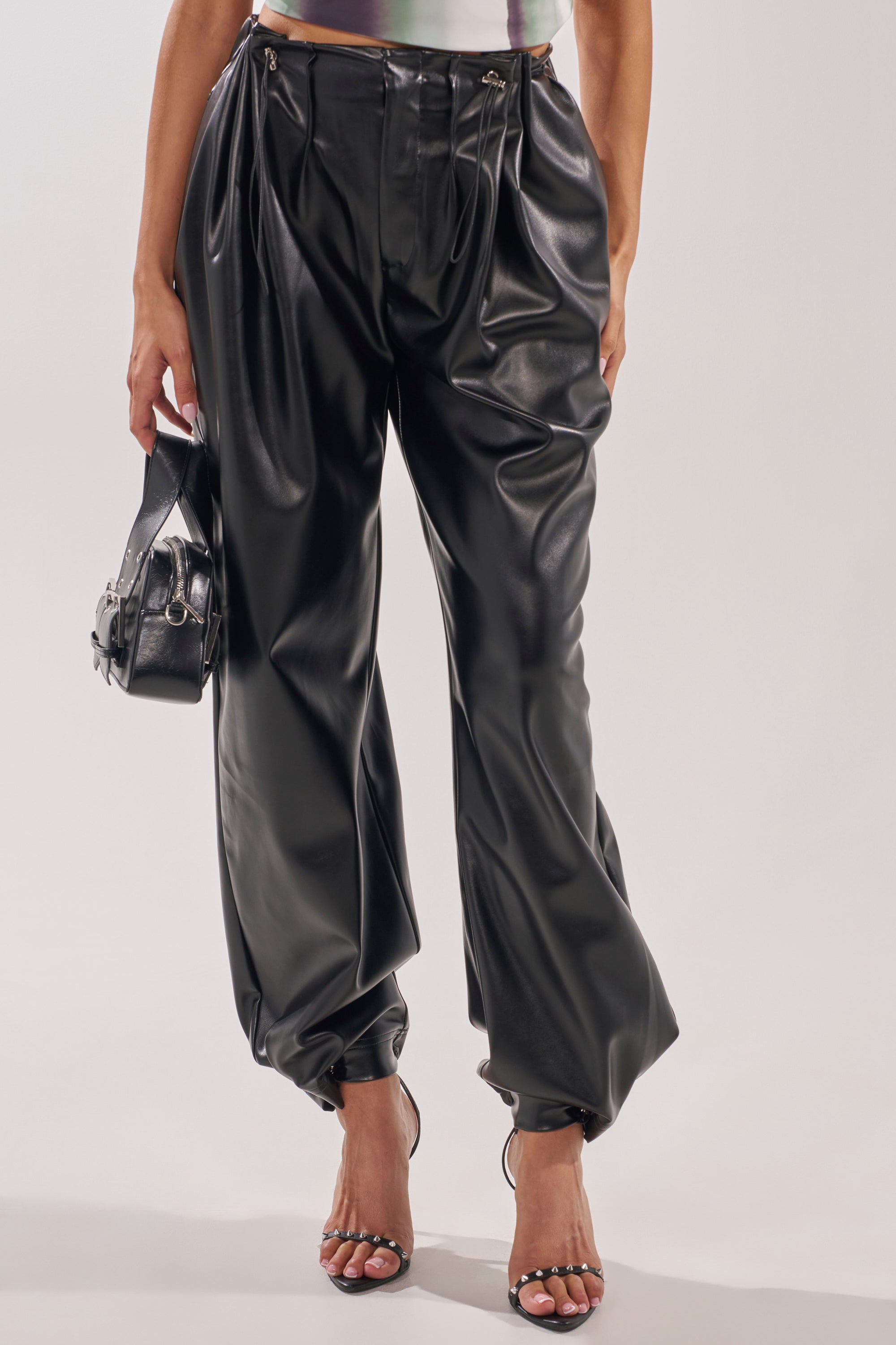 A person wears the ON TOP OF IT FAUX LEATHER TROUSER—high-rise, wide-leg with pleats—paired with a small black handbag and strappy heeled sandals against a plain, light background. 2