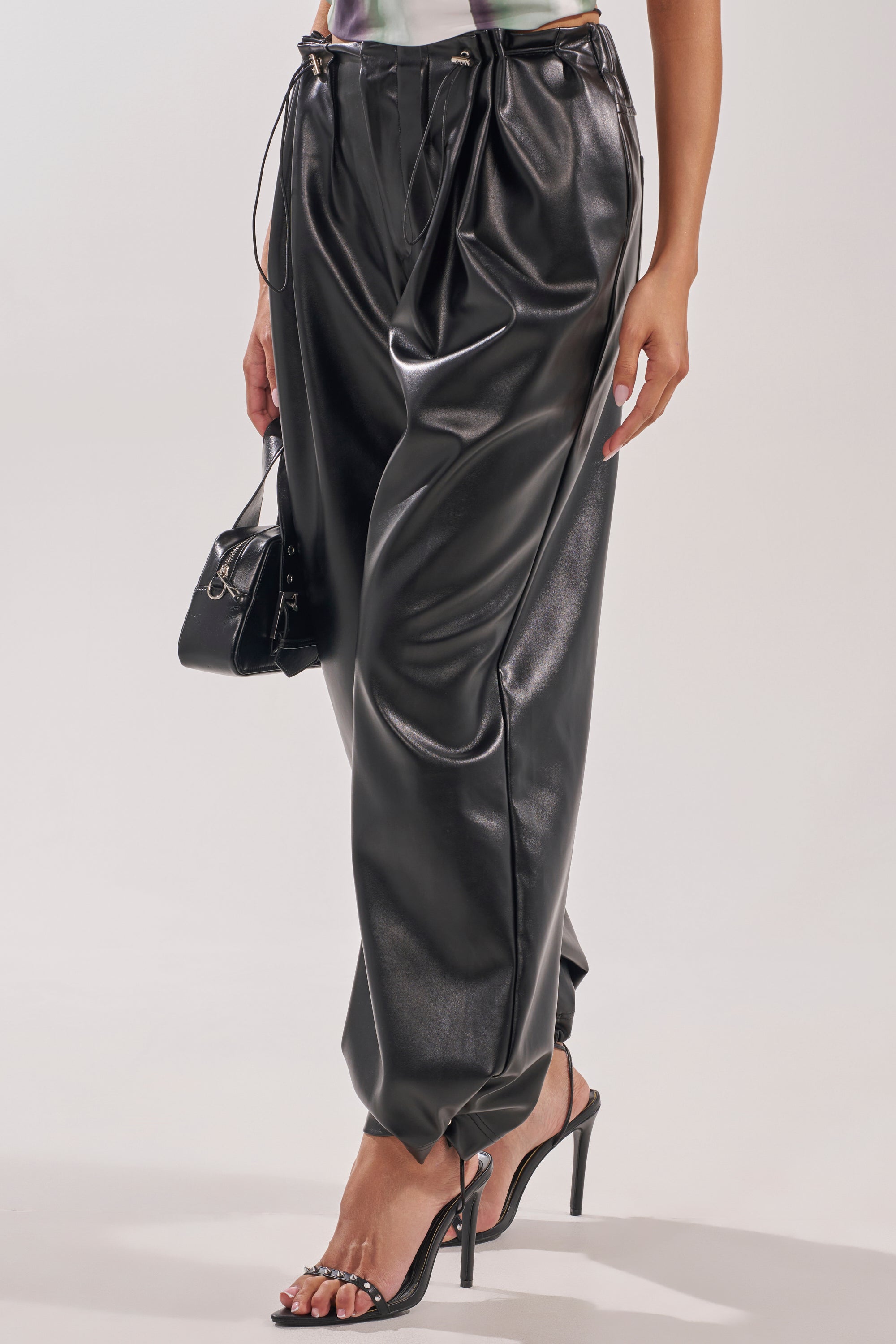 A person models the ON TOP OF IT FAUX LEATHER TROUSER—high-rise, baggy faux leather pants with drawstring ankles—paired with a small black handbag and black high-heeled sandals against a plain background. 3