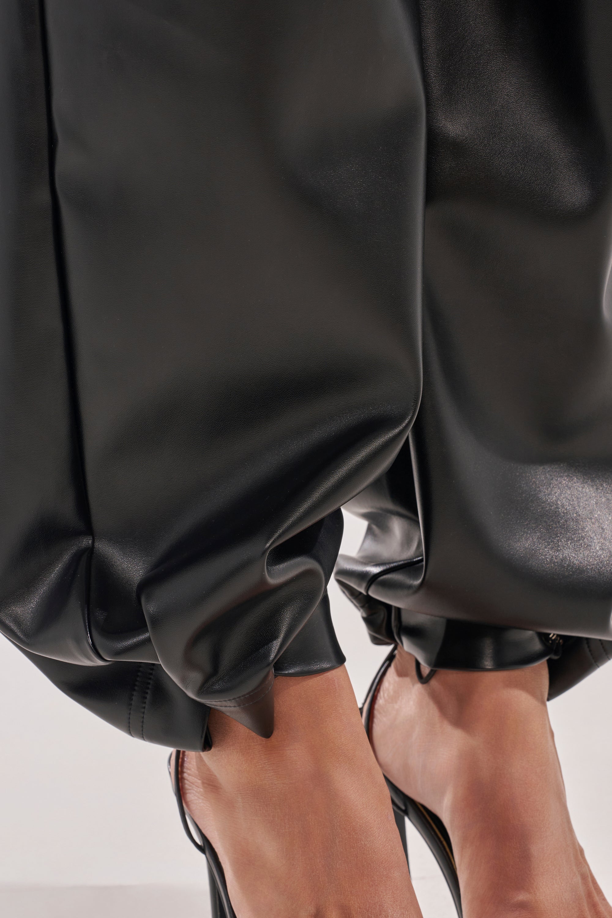 Close-up of a person wearing ON TOP OF IT FAUX LEATHER TROUSER, featuring shiny black high-rise fit and gathered ankle cuffs, paired with clear high-heeled shoes that show their toes against a plain light background. 7