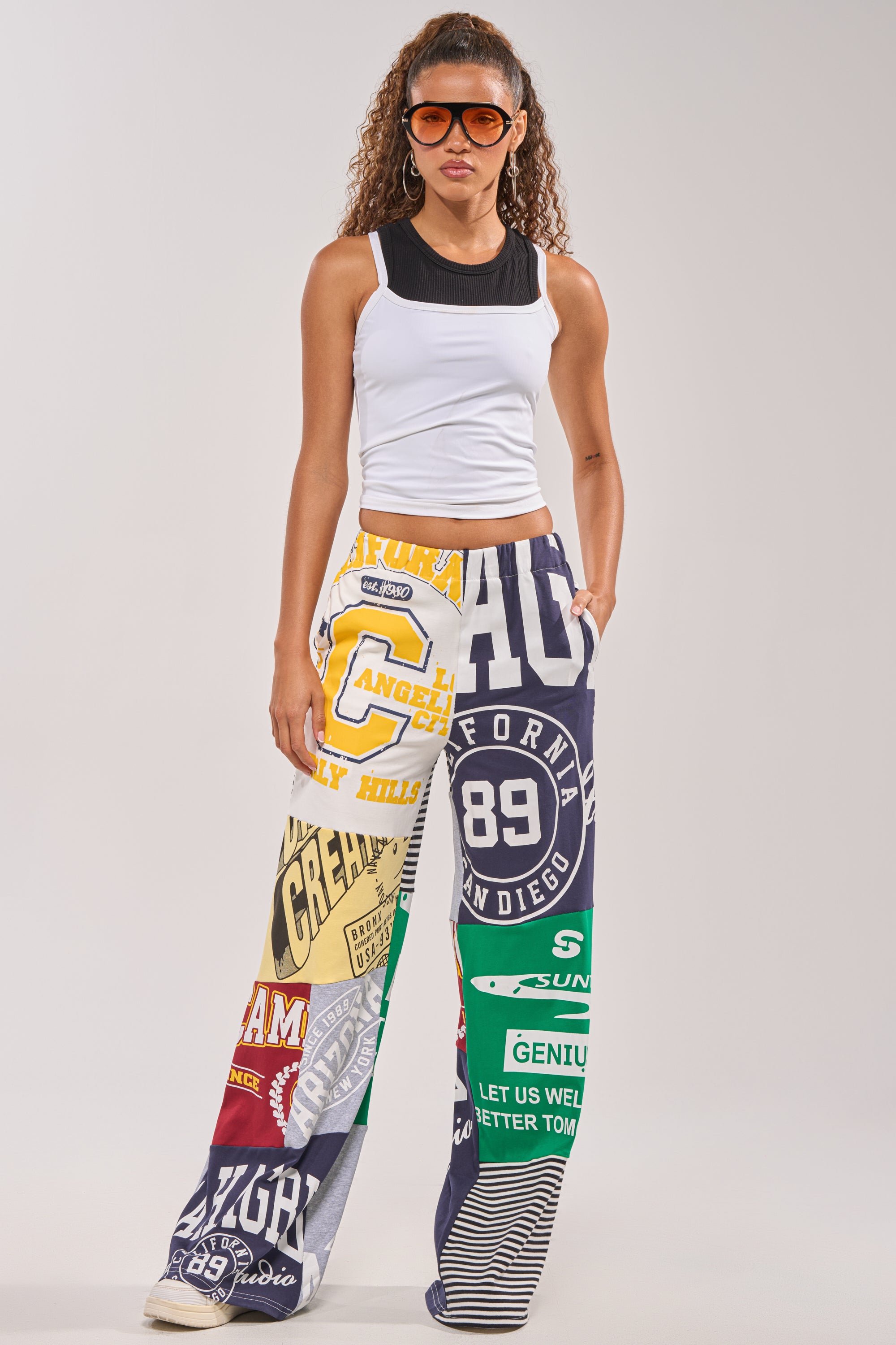 A woman poses against a plain background wearing large sunglasses, a sleeveless white top, and the GAME TIME PATCHWORK JOGGER featuring colorful streetwear-inspired designs with bold logos, numbers, and graphic text. 0