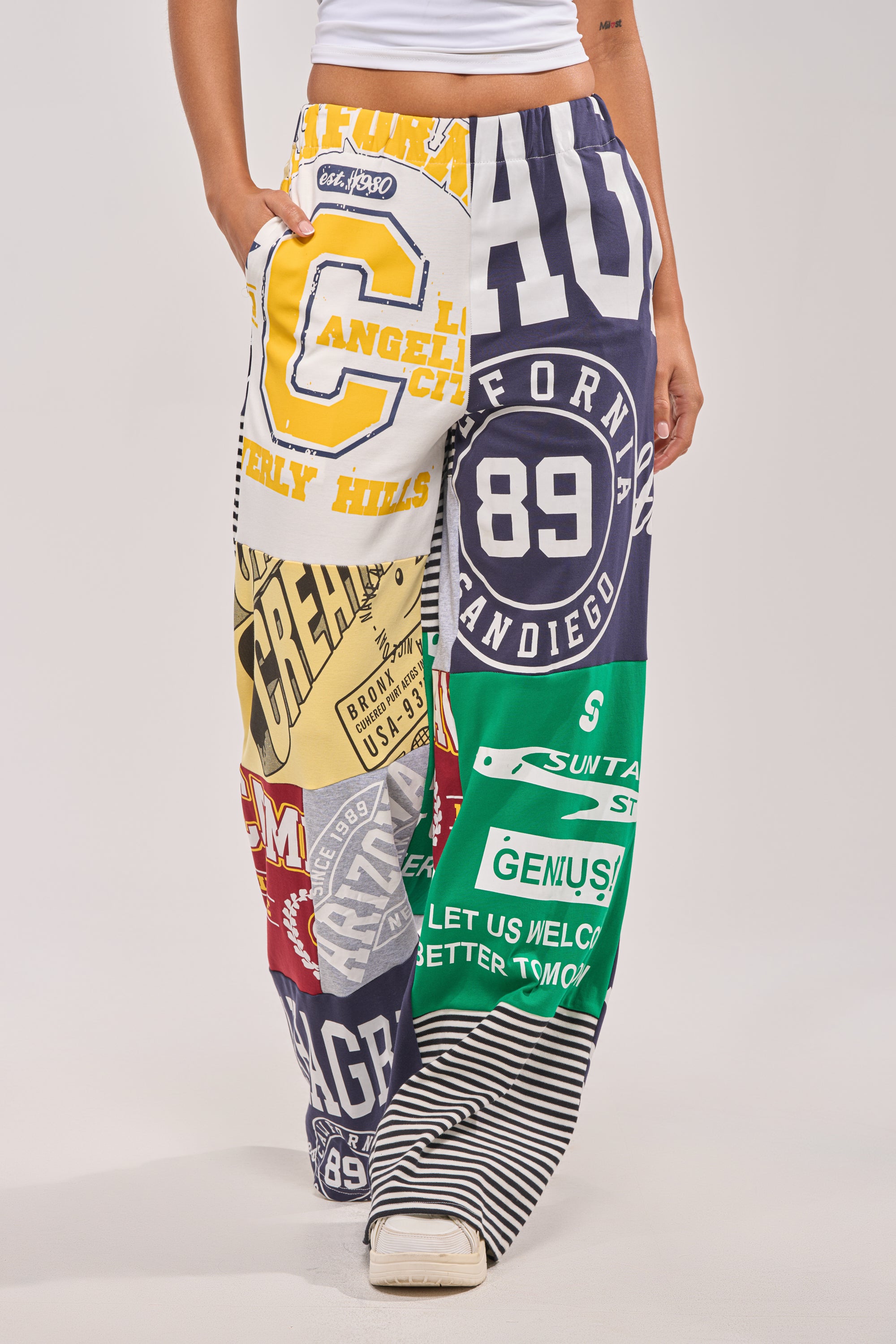Someone wears the GAME TIME PATCHWORK JOGGER, featuring bold graphics and logos in yellow, green, red, black, and white, styled with white sneakers and a white top for a cool streetwear look. 1