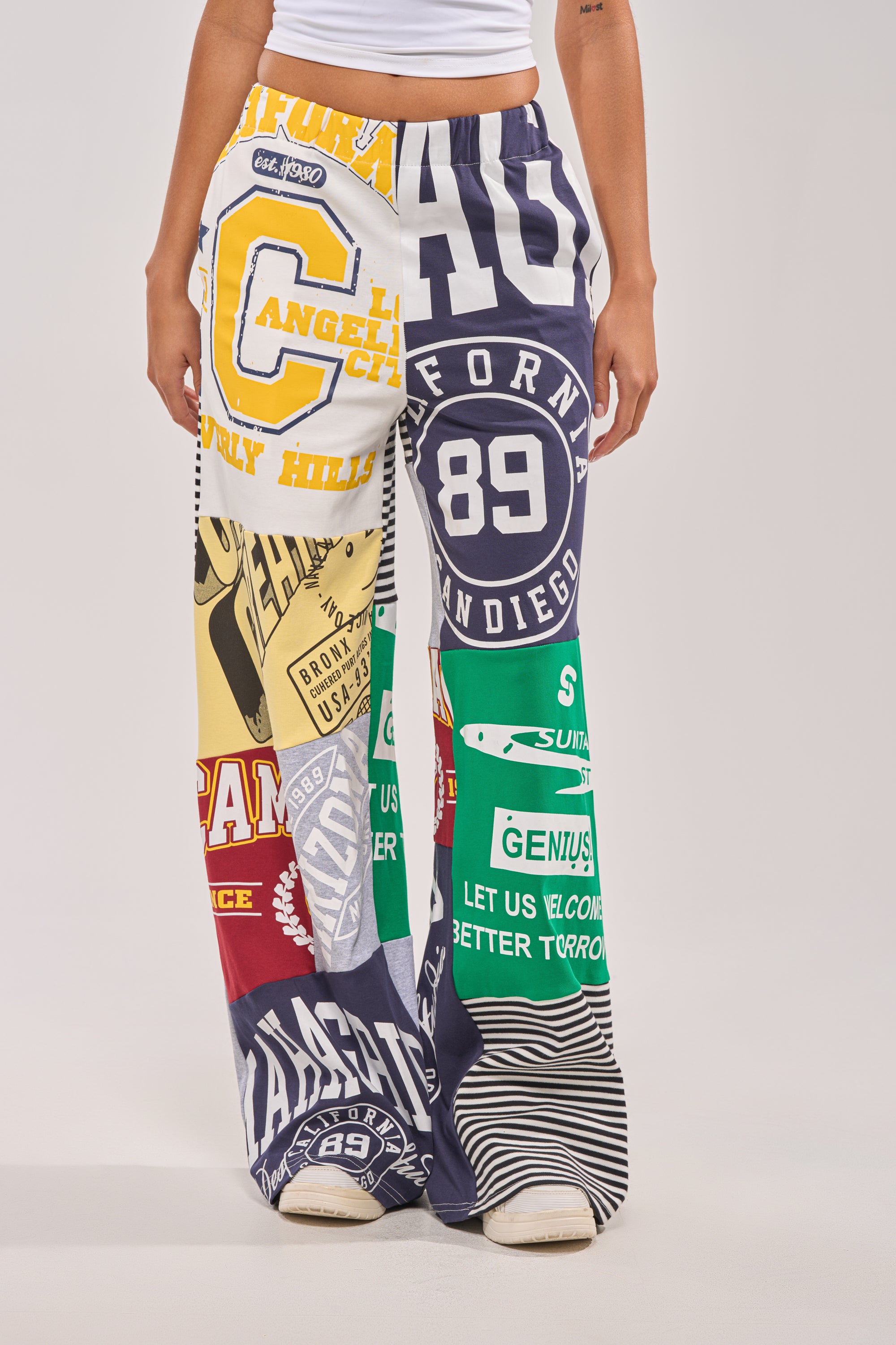 A person models the GAME TIME PATCHWORK JOGGER, featuring bold college-style graphics, numbers, and text patches in a colorful design. Only their torso and legs are visible against a plain light background. 2