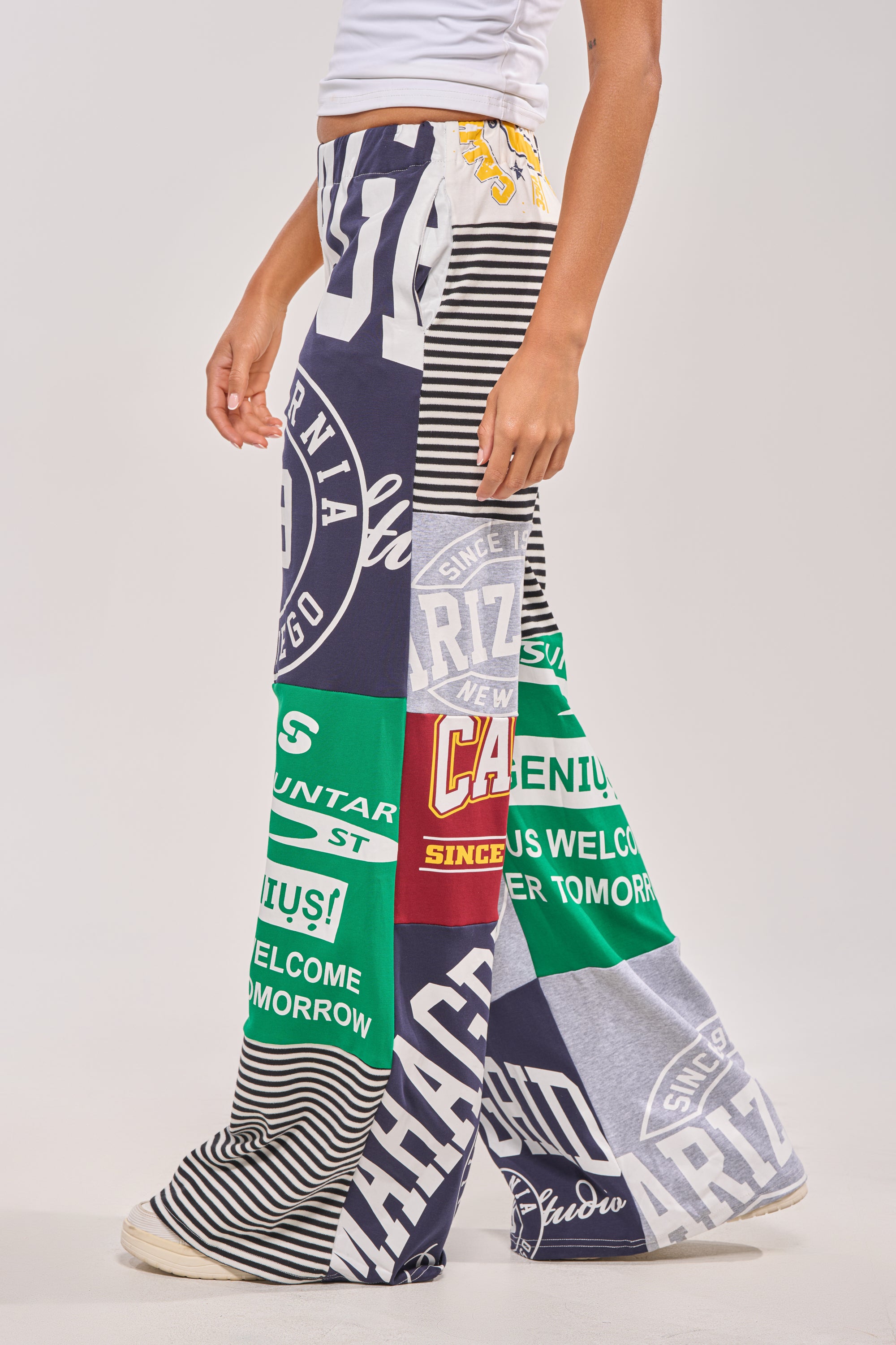 A person wears GAME TIME PATCHWORK JOGGER pants with colorful graphic prints, stripes, and bold logos. These streetwear joggers have a relaxed fit, paired with white sneakers against a plain light background. 3