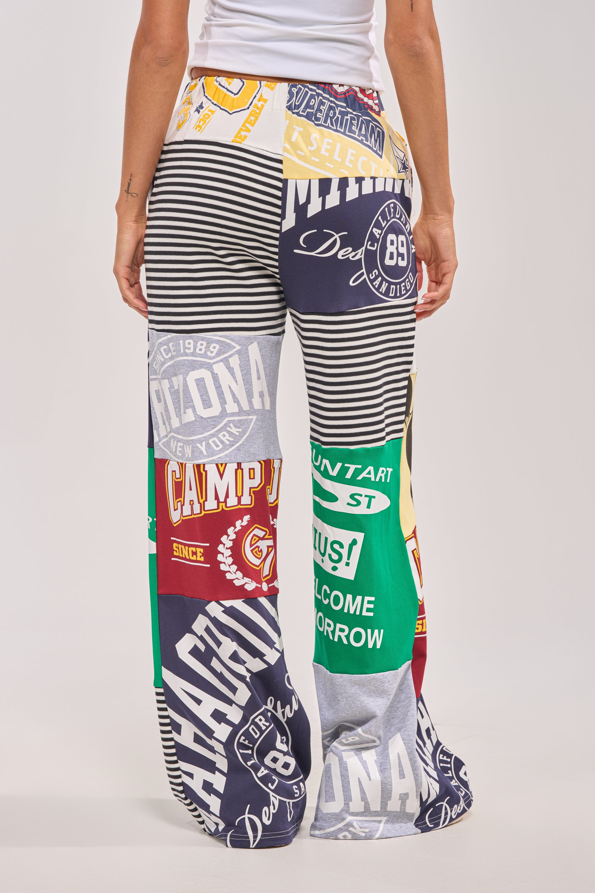 Someone is seen from behind wearing GAME TIME PATCHWORK JOGGER—high-waisted, wide-leg joggers featuring bold stripes, college-themed graphics, text, and vibrant patterns—against a plain background. 5