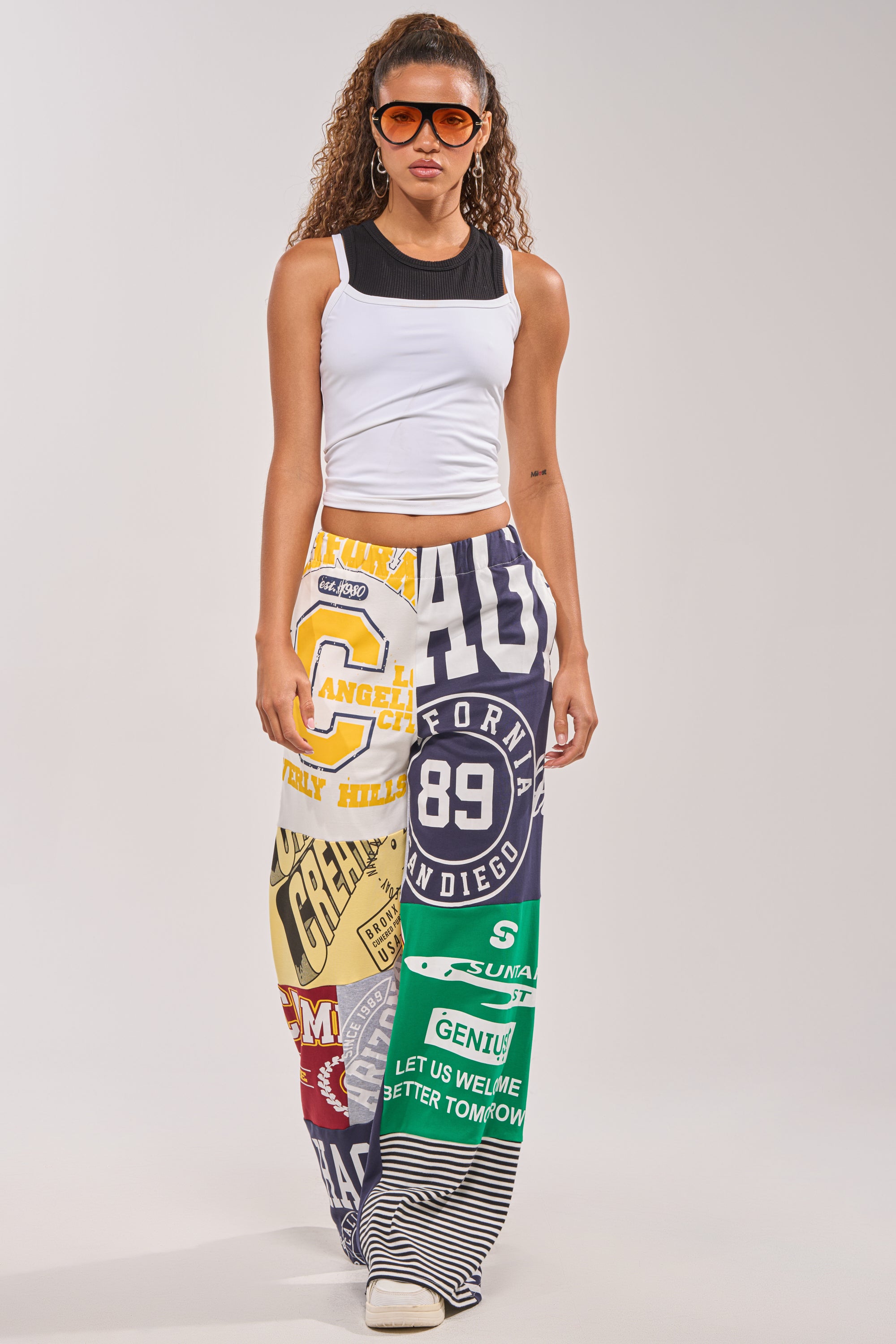 A woman with long curly hair and orange sunglasses wears a white crop top with black trim and the GAME TIME PATCHWORK JOGGER—streetwear-inspired, wide-leg pants featuring colorful, bold graphic prints and logos—against a light background. 6