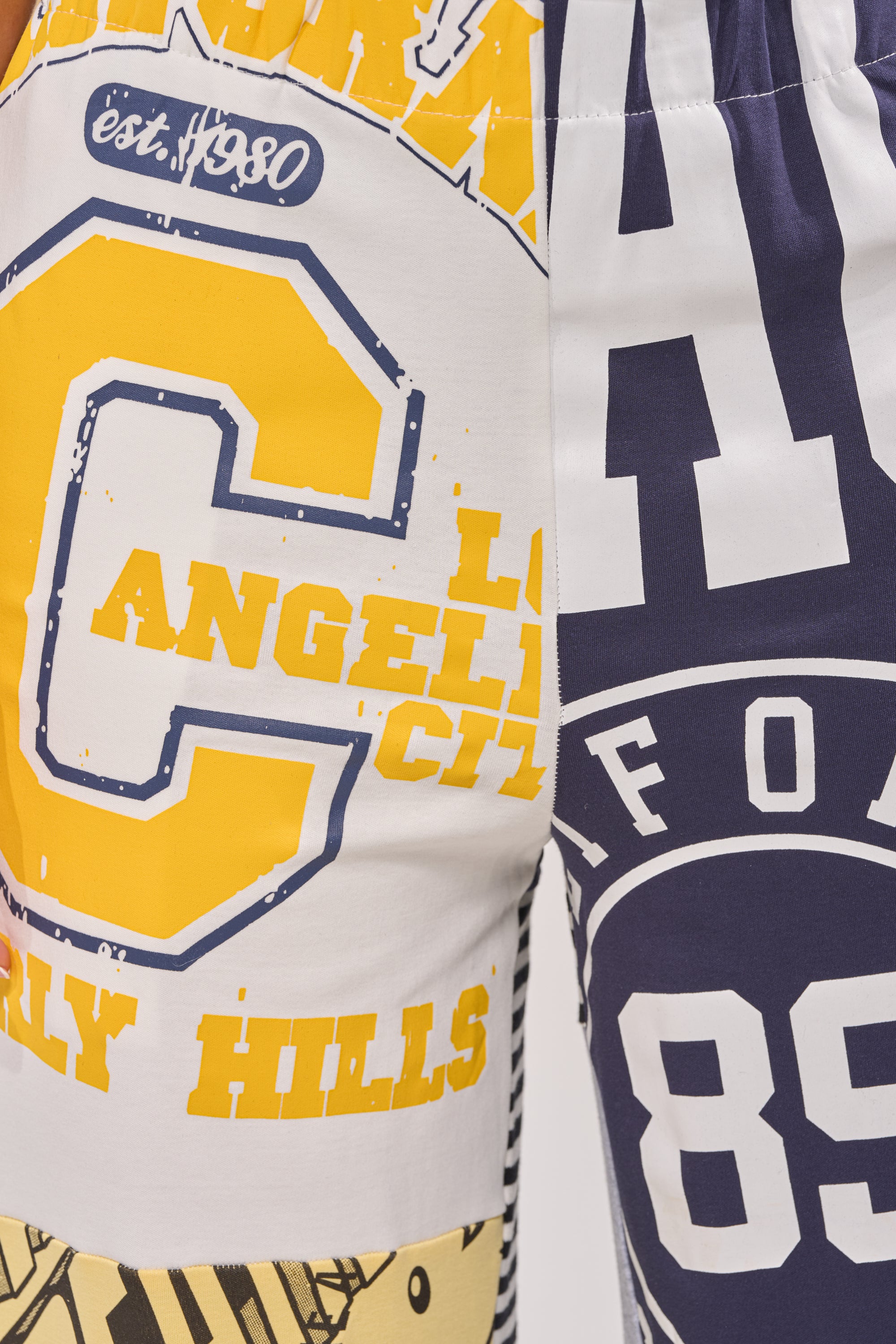 A close-up of the GAME TIME PATCHWORK JOGGER shows two-toned streetwear shorts with bold yellow and navy blue collegiate graphics, including "Los Angeles," "Hills," "est. 1980," and the number "89. 7