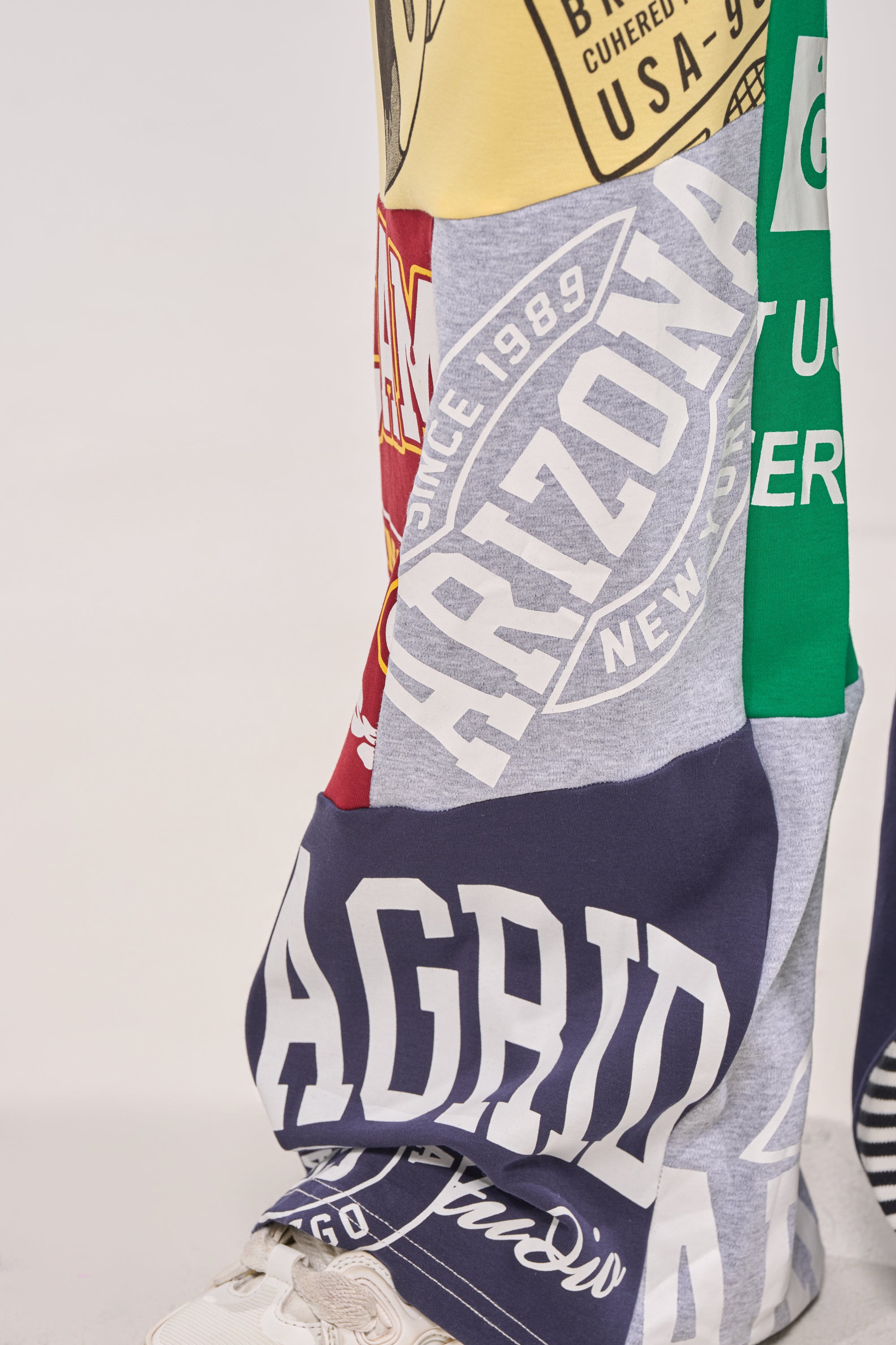 A close-up of a person wearing the GAME TIME PATCHWORK JOGGER, crafted from colorful graphic T-shirt fabrics with words like "Arizona" and "Madrid." The wide-leg joggers are paired with white sneakers against a plain light background. 8