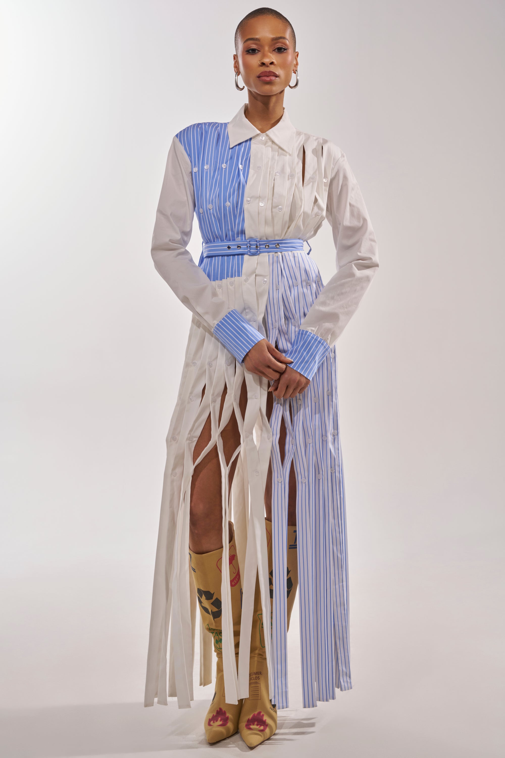 A woman models the NOT MY MANS BUTTON UP SHIRT MAXI DRESS with vertical blue stripes and fringe accents, styled with tall tan floral boots, statement earrings, and a confident look against a plain background. 0