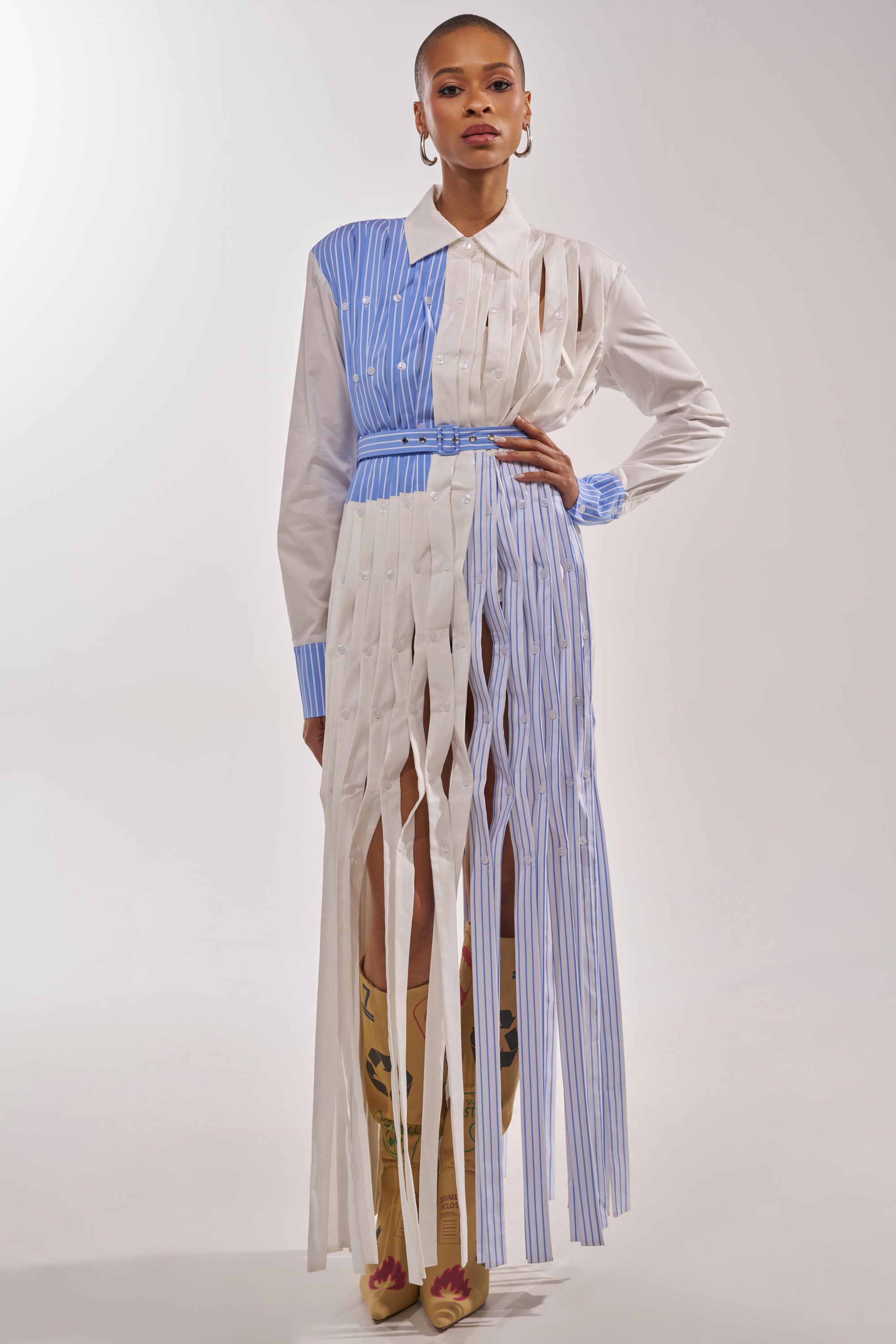 A model wears the NOT MY MANS BUTTON UP SHIRT MAXI DRESS with alternating white and blue pinstripes and vertical fringe-like cuts, styled with a belt, hoop earrings, and patterned tan boots for a chic business casual look. 1
