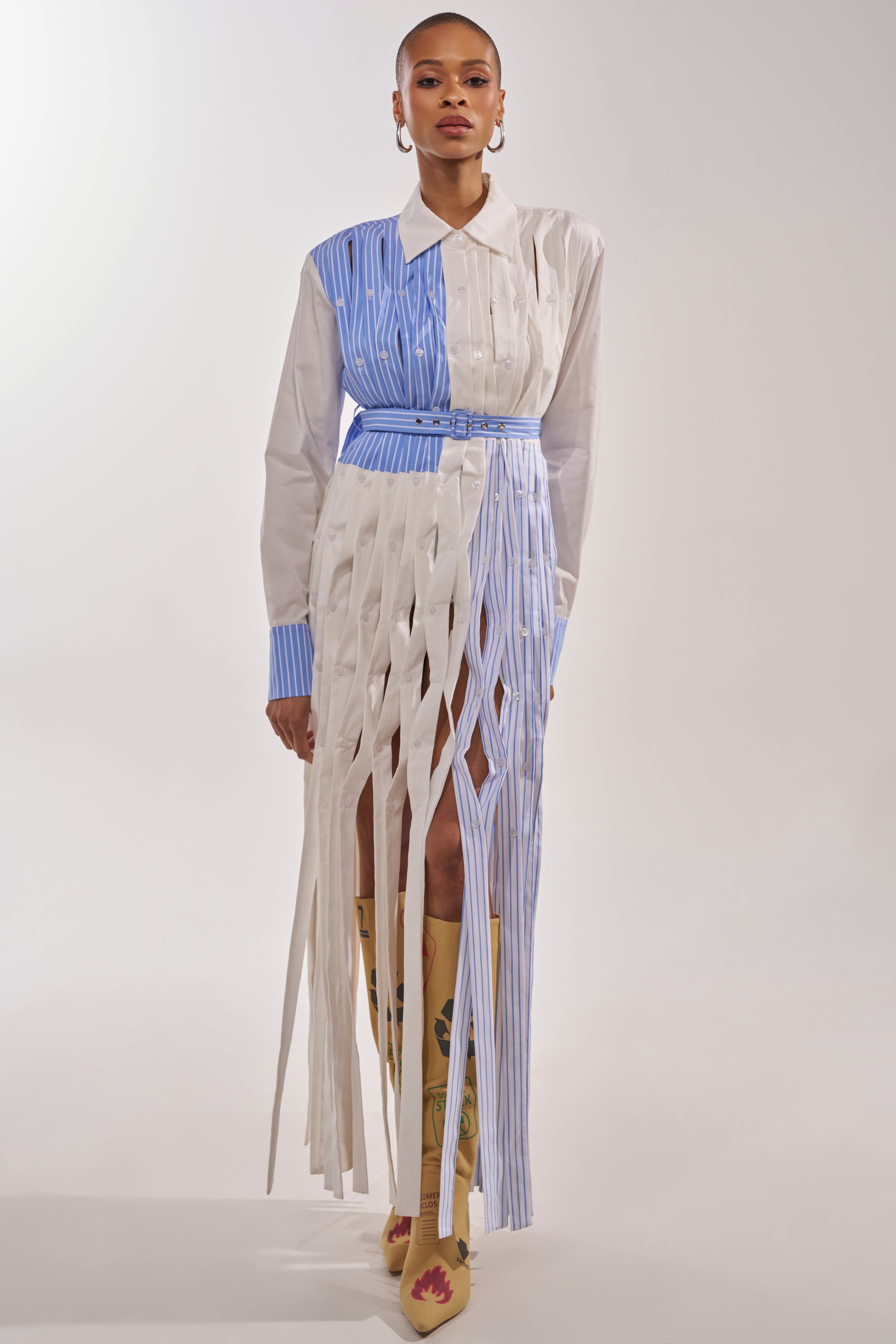 A person wears the NOT MY MANS BUTTON UP SHIRT MAXI DRESS, a long-sleeve blue and white striped shirt dress with a fringe skirt, styled with tall tan boots featuring blue and red designs, against a plain white background. 2