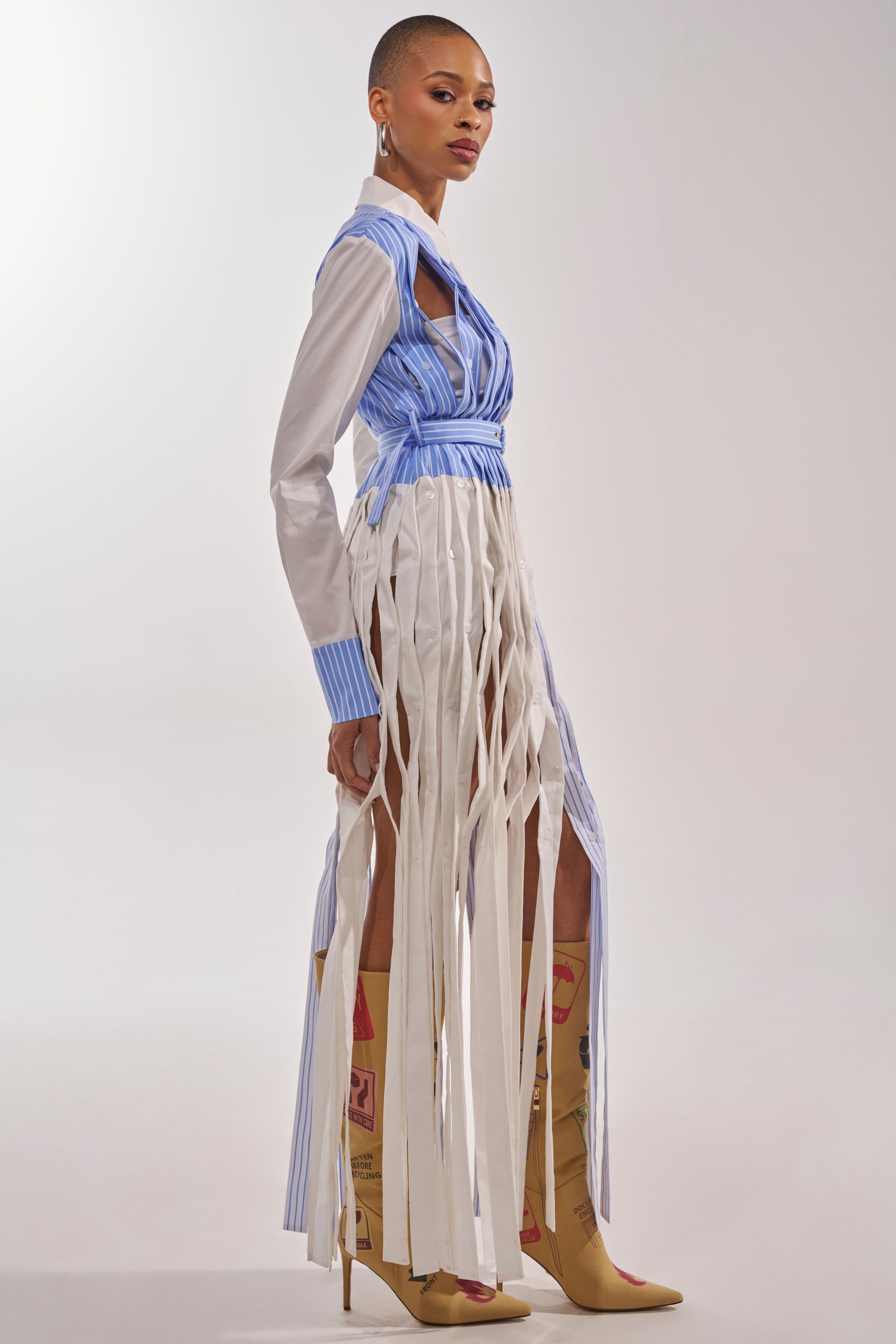 A person stands in profile wearing the NOT MY MANS BUTTON UP SHIRT MAXI DRESS, a blue and white striped panel dress with long vertical fringes, paired with tall tan boots featuring red and yellow designs against a plain light background. 3