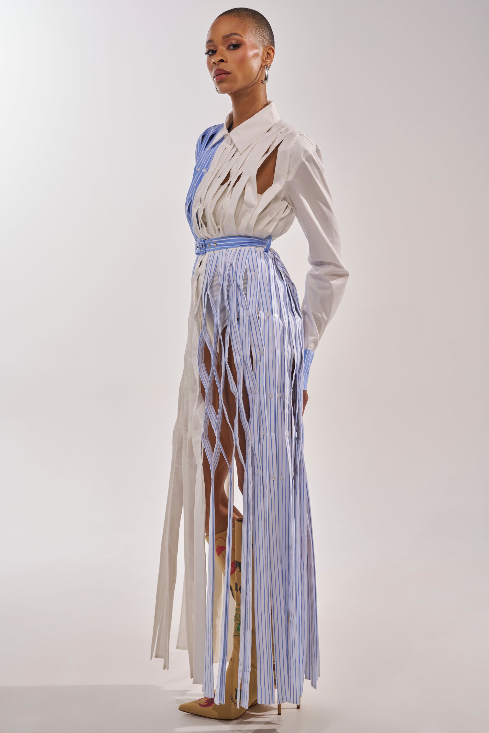 A model with a shaved head wears the NOT MY MANS BUTTON UP SHIRT MAXI DRESS, featuring vertical white and blue strips for a fringe effect, paired with tall pointed-toe boots against a simple backdrop. 4