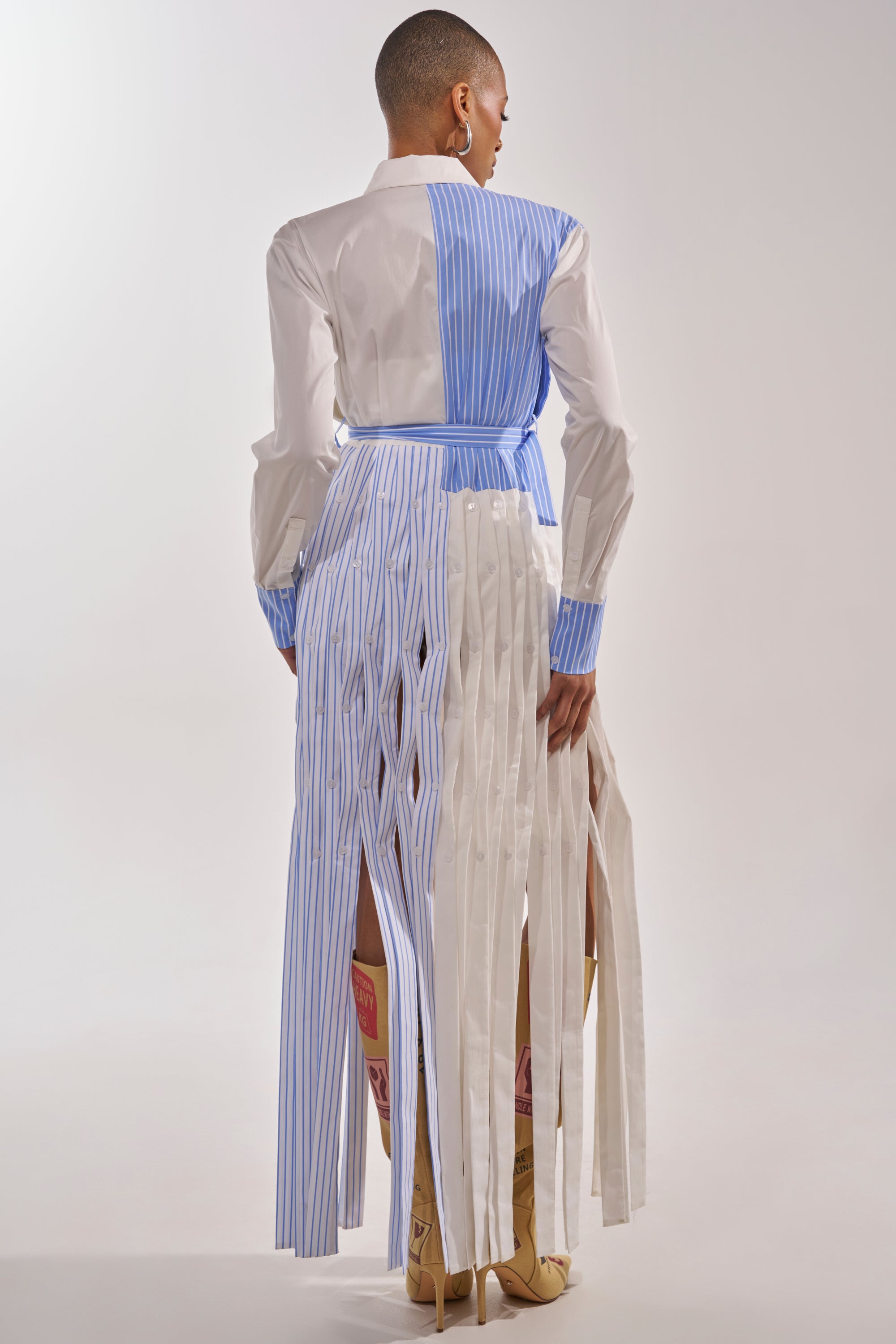 A person faces away, wearing the NOT MY MANS BUTTON UP SHIRT MAXI DRESS with vertical blue and white stripes. The skirt features slatted, crisscross panels revealing tall tan boots against a plain, light background. 5