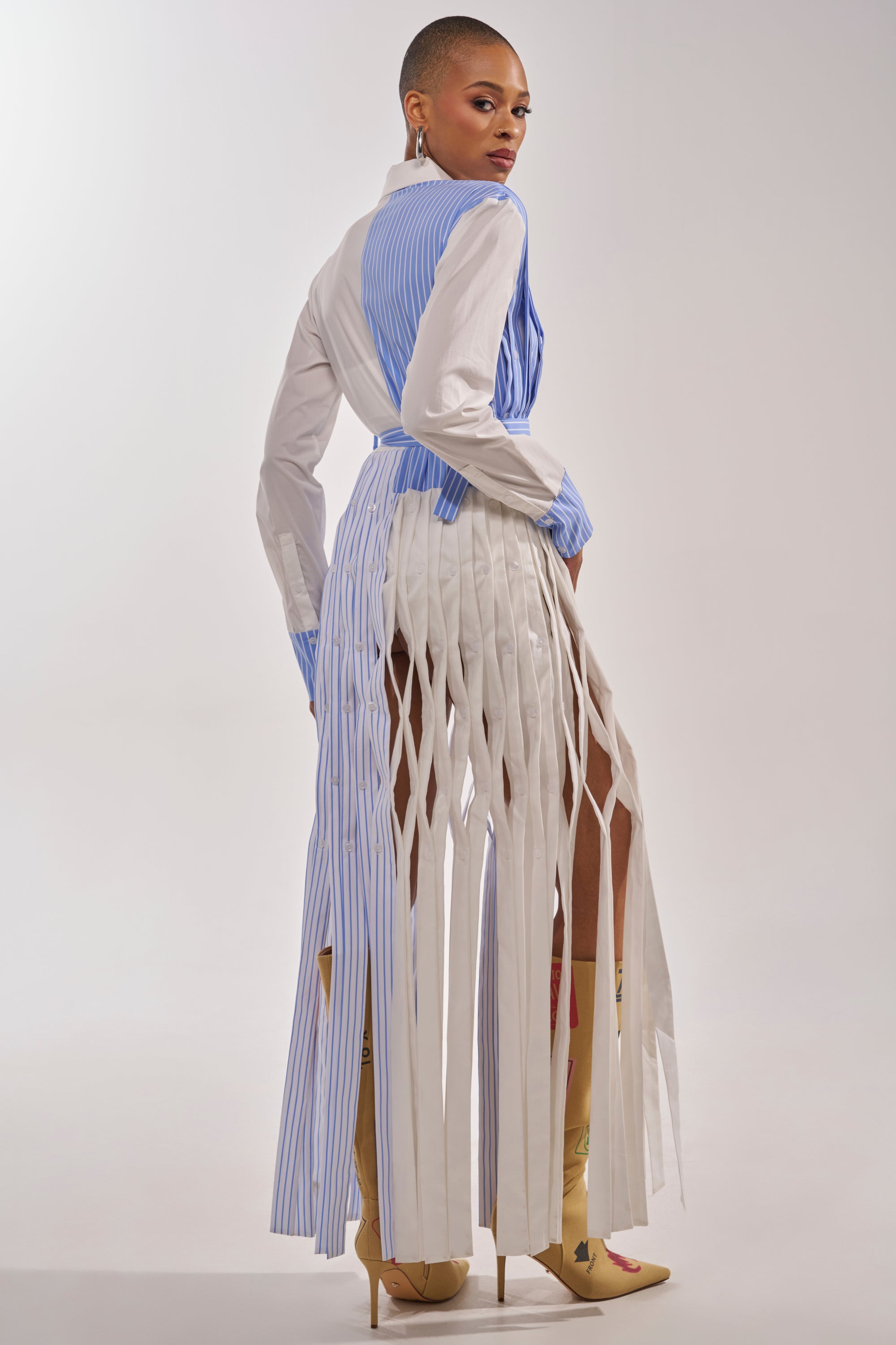 A woman with short hair poses in the NOT MY MANS BUTTON UP SHIRT MAXI DRESS, featuring striped panels and vertical fringe cuts. She pairs it with tall beige stiletto boots against a plain, light-colored background. 6