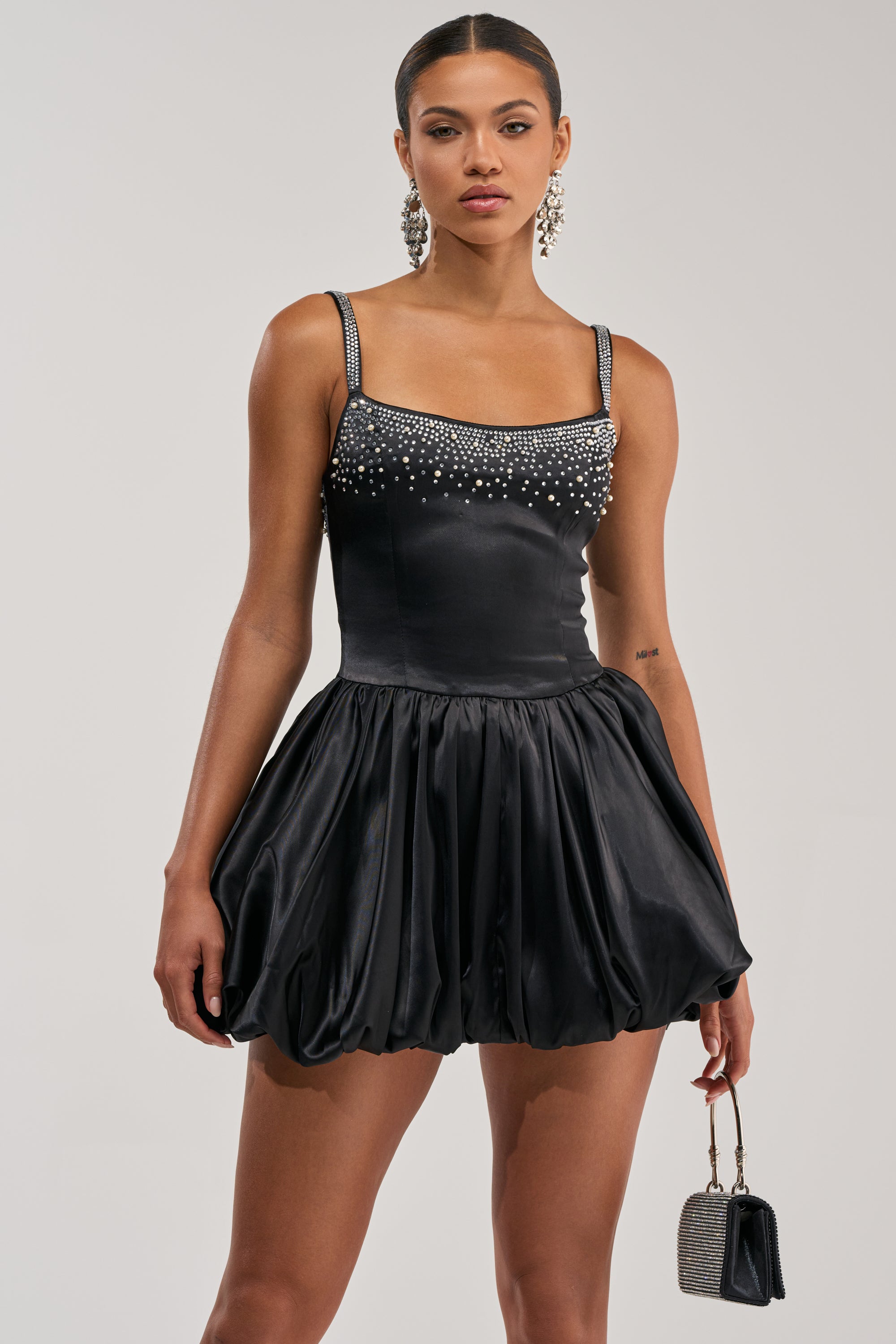 A woman in the COSMIC DREAM EMBELLISHED MINI DRESS, featuring a sleeveless black satin design with a beaded neckline and bubble skirt, holds a silver handbag. She styles her slicked-back hair with large dangling earrings.