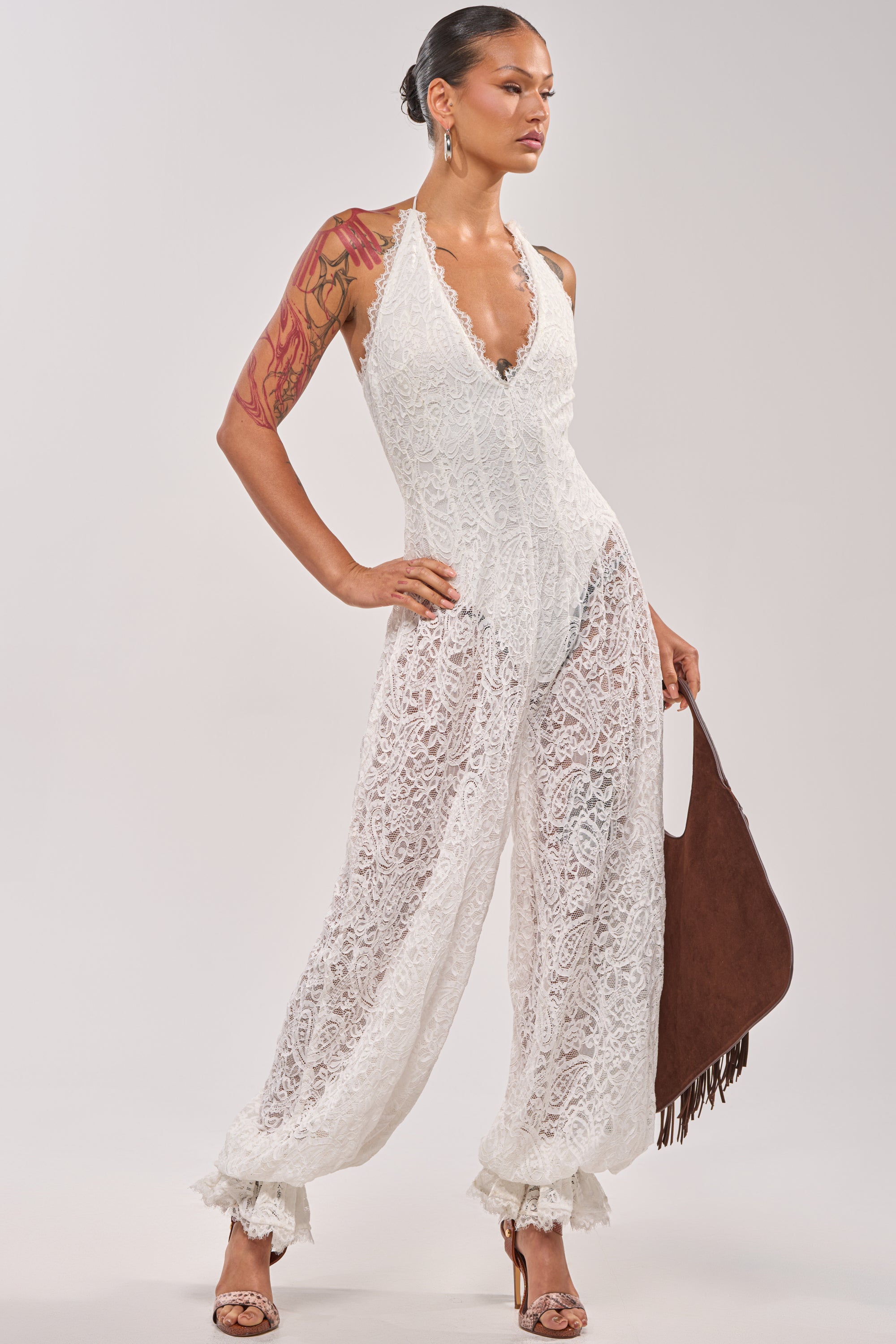 A woman models the GALAVANTING LACE JUMPSUIT IN IVORY, a sleeveless piece with ruffled cuffs. She accessorizes with hoop earrings and a brown fringed bag, her tattoos visible and hair pulled back—a chic summer look. 3