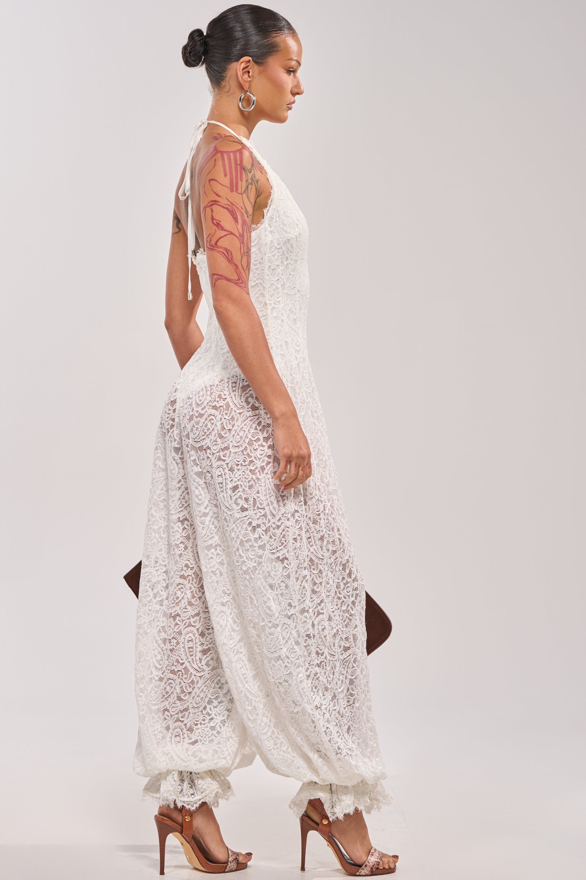A woman in profile wears the GALAVANTING LACE JUMPSUIT IN IVORY, a sleeveless white halter style with loose wide legs and fitted cuffs. She pairs it with high heels, hoop earrings, an updo, a brown clutch, and displays arm tattoos. 4