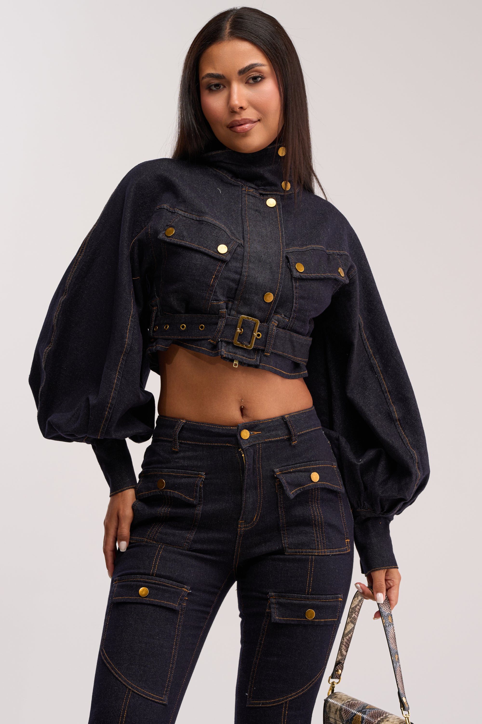 A woman poses confidently in the CHIARA DENIM JACKET—a utility-inspired, cropped denim jacket with balloon sleeves, gold buttons, and a belt—paired with matching high-waisted jeans. She carries a small patterned handbag and smiles slightly.
