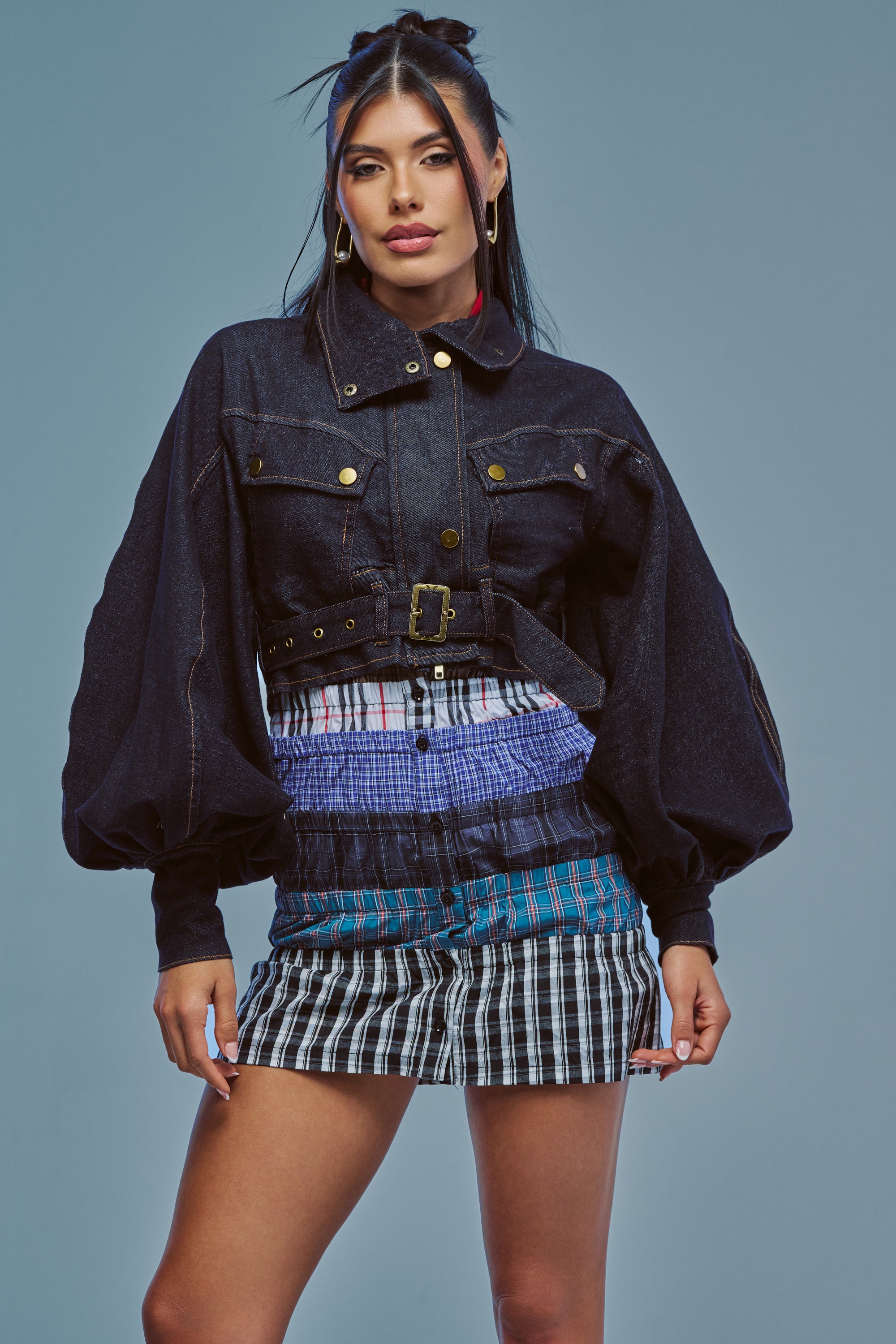 A woman with long dark hair and gold hoop earrings poses against a blue background wearing the CHIARA DENIM JACKET, featuring puffy sleeves and a belted waist, paired with a short plaid skirt in mixed colors.