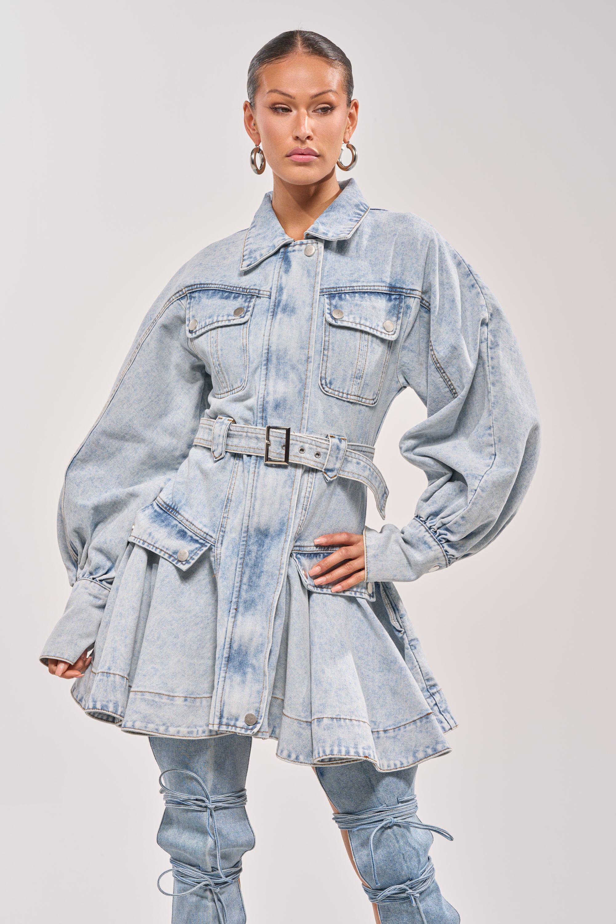 A woman models the SAY PRETTY PLEASE DENIM MINI DRESS, featuring a belted waist, oversized sleeves, and front pockets. She pairs it with large hoop earrings and pulled-back hair for a chic update on the classic denim dress look. 0
