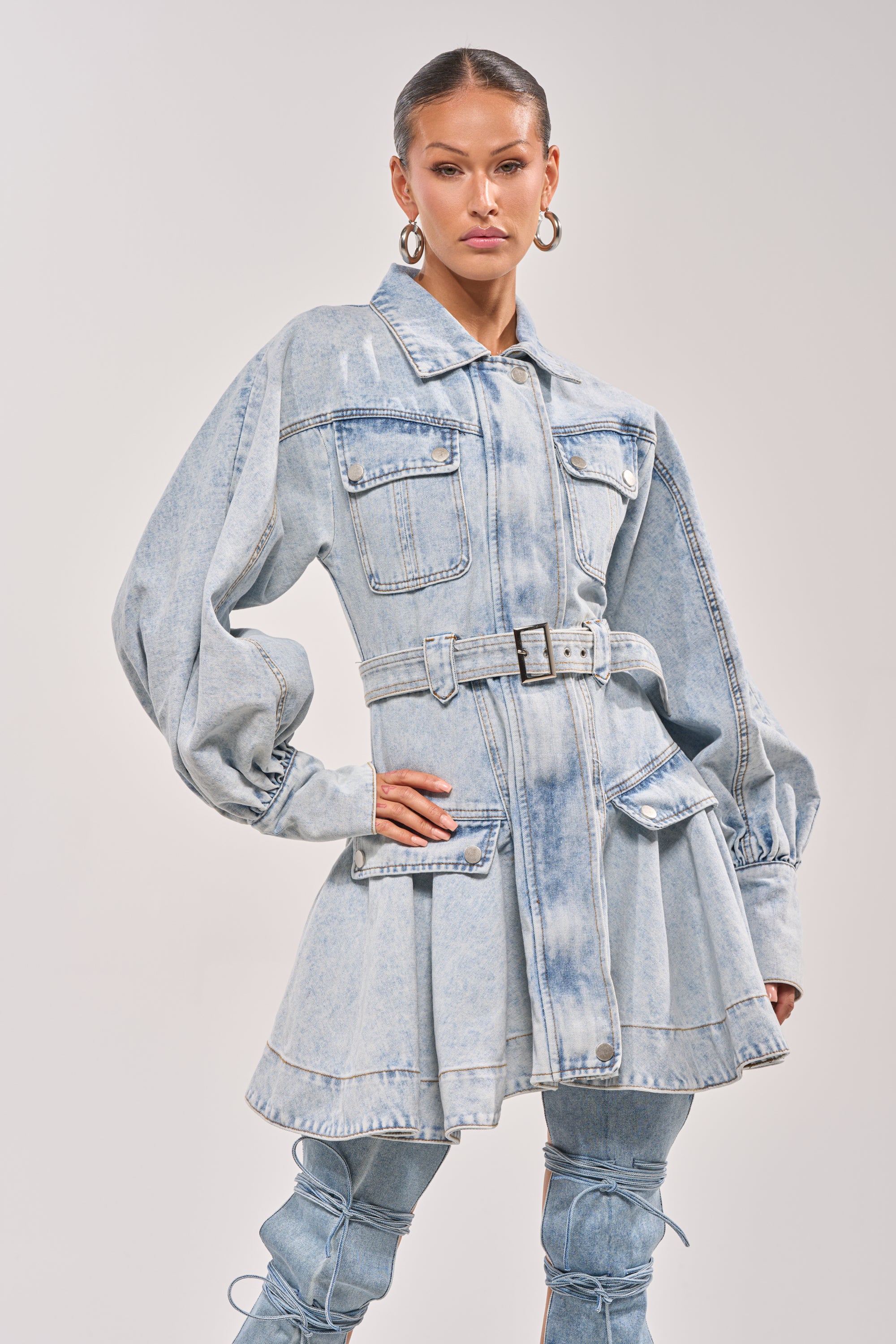 Someone poses confidently in the SAY PRETTY PLEASE DENIM MINI DRESS featuring a belted fit and flare silhouette, puffed sleeves, and large front pockets, styled with matching denim pants and hoop earrings against a plain background. 1