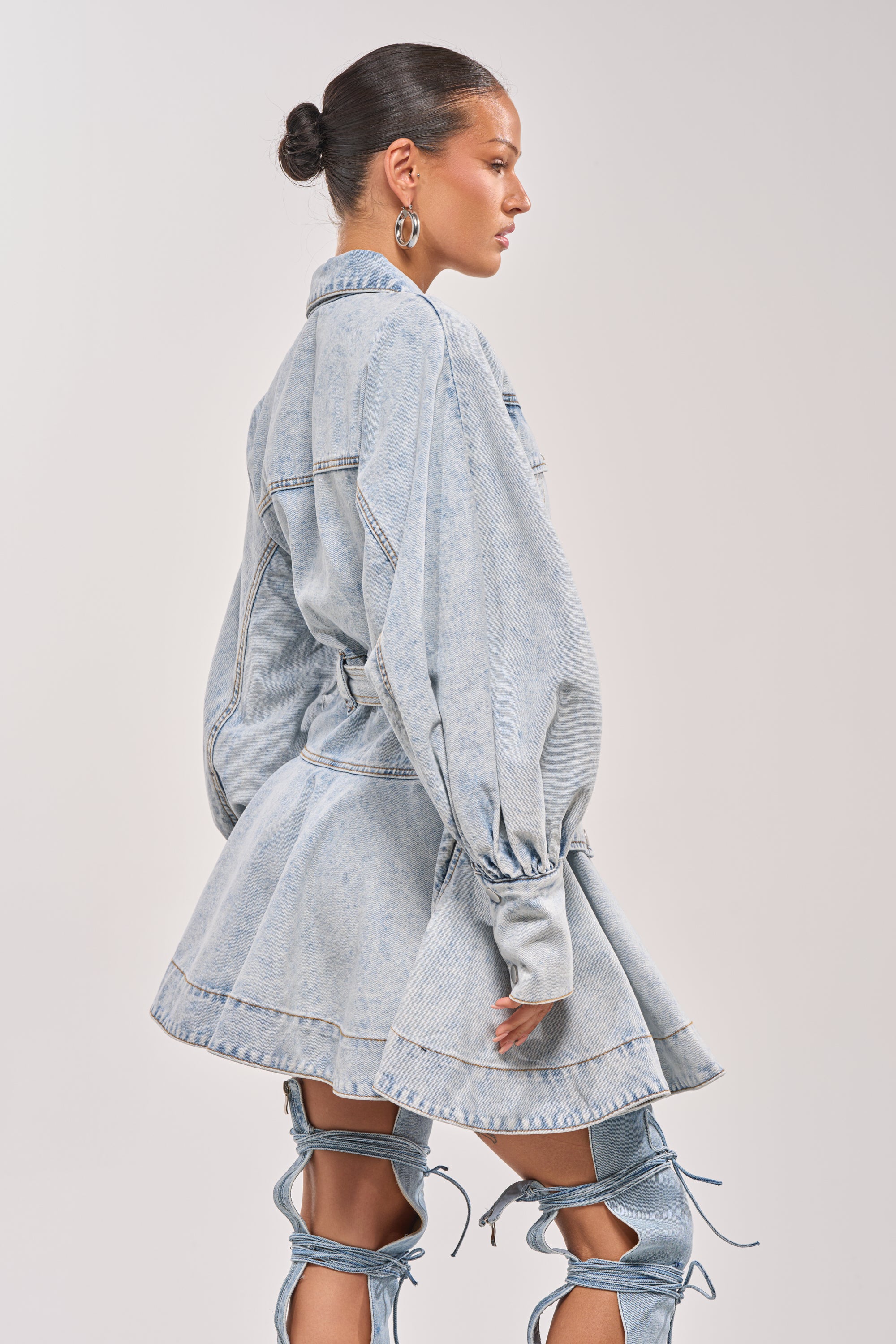A woman stands in profile against a plain background wearing the SAY PRETTY PLEASE DENIM MINI DRESS with a belted waist, puffed sleeves, flared skirt, and matching over-the-knee lace-up boots. 2