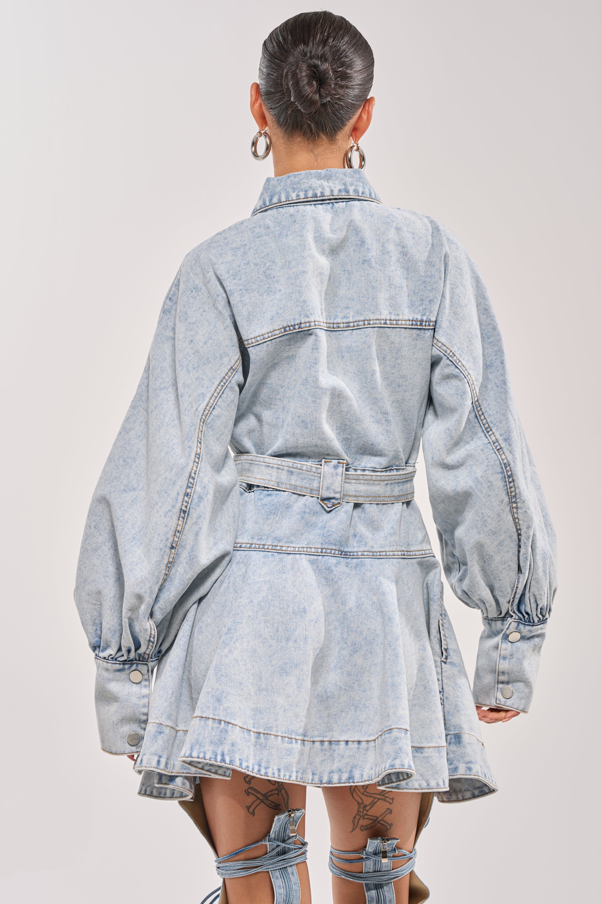 A person with their hair in a bun wears the SAY PRETTY PLEASE DENIM MINI DRESS, a light blue belted mini dress with long, puffed sleeves, large cuffs, visible seams, and a flared skirt as they face away from the camera. 3