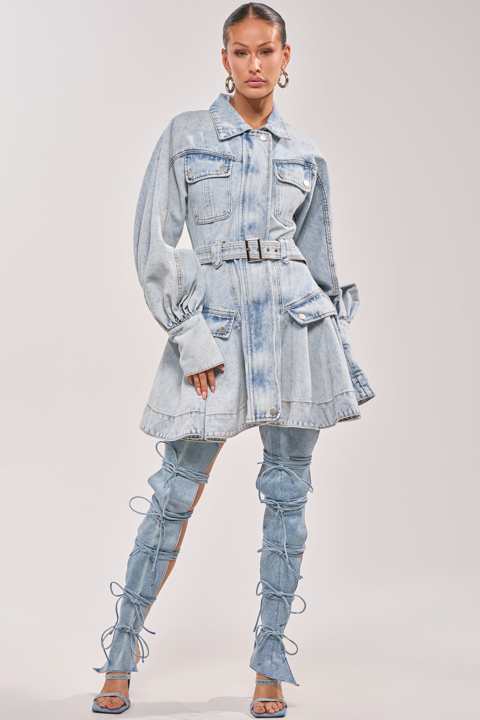 A woman poses confidently in the SAY PRETTY PLEASE DENIM MINI DRESS, featuring a belted waist, long sleeves, oversized pockets, and matching lace-up thigh-high denim boots against a plain light background. 4