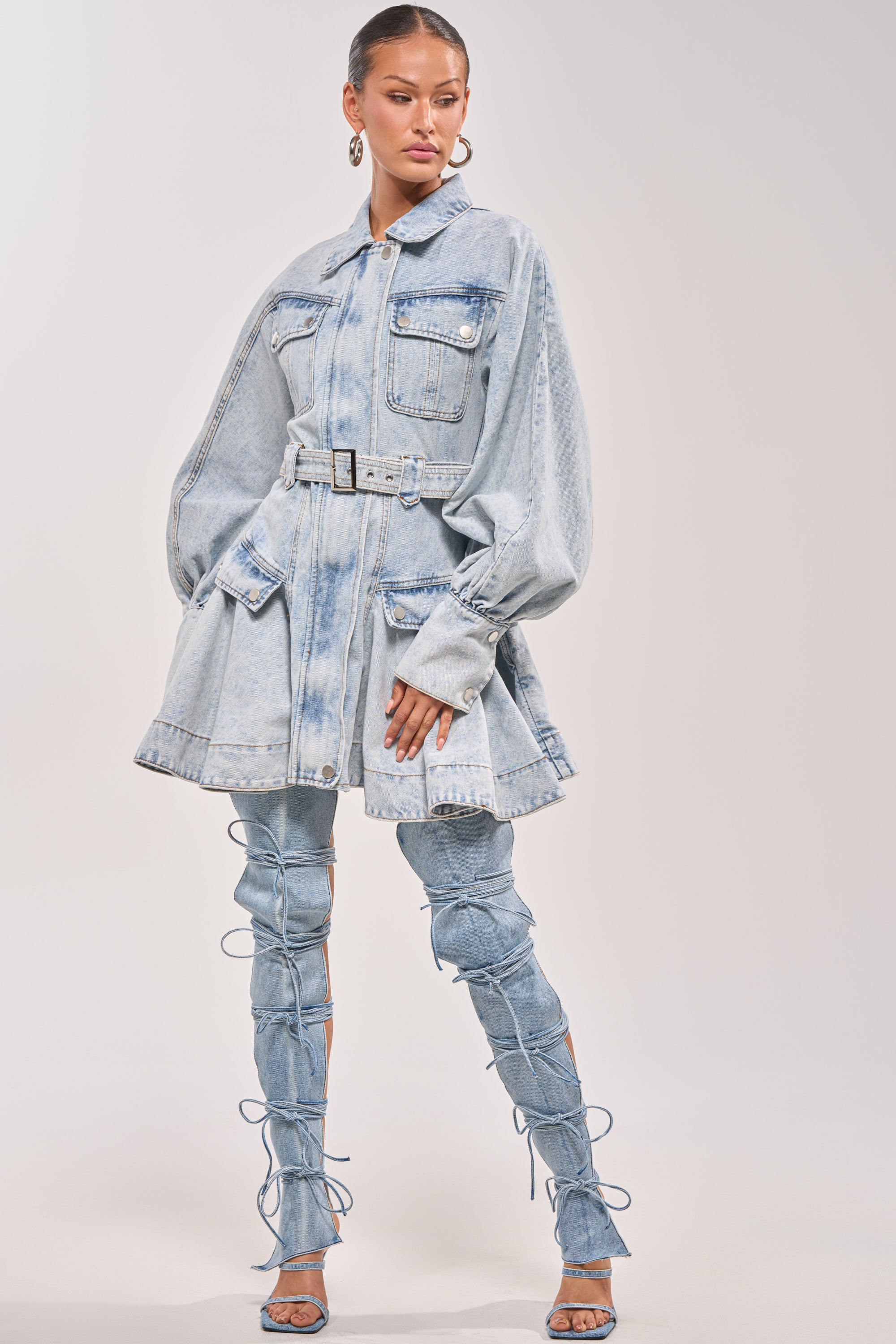 A woman models the SAY PRETTY PLEASE DENIM MINI DRESS, featuring a belted waist, oversized sleeves, and large front pockets, paired with matching lace-up high-heeled denim boots against a plain light background. 5