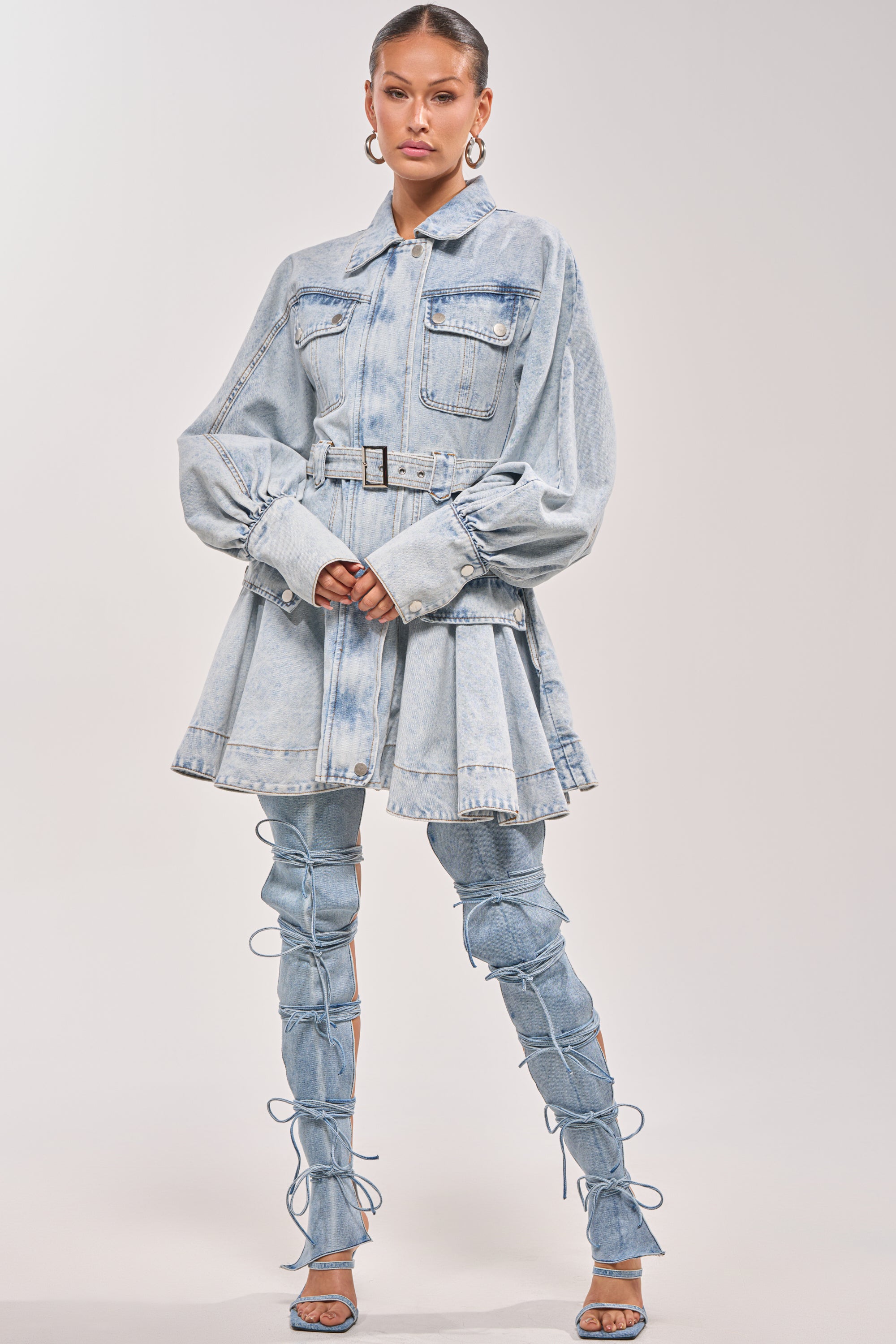 A woman poses in the SAY PRETTY PLEASE DENIM MINI DRESS, styled with puffed sleeves, a belted waist, matching lace-up thigh-high denim boots with high heels, hoop earrings, and her hair pulled back. 6