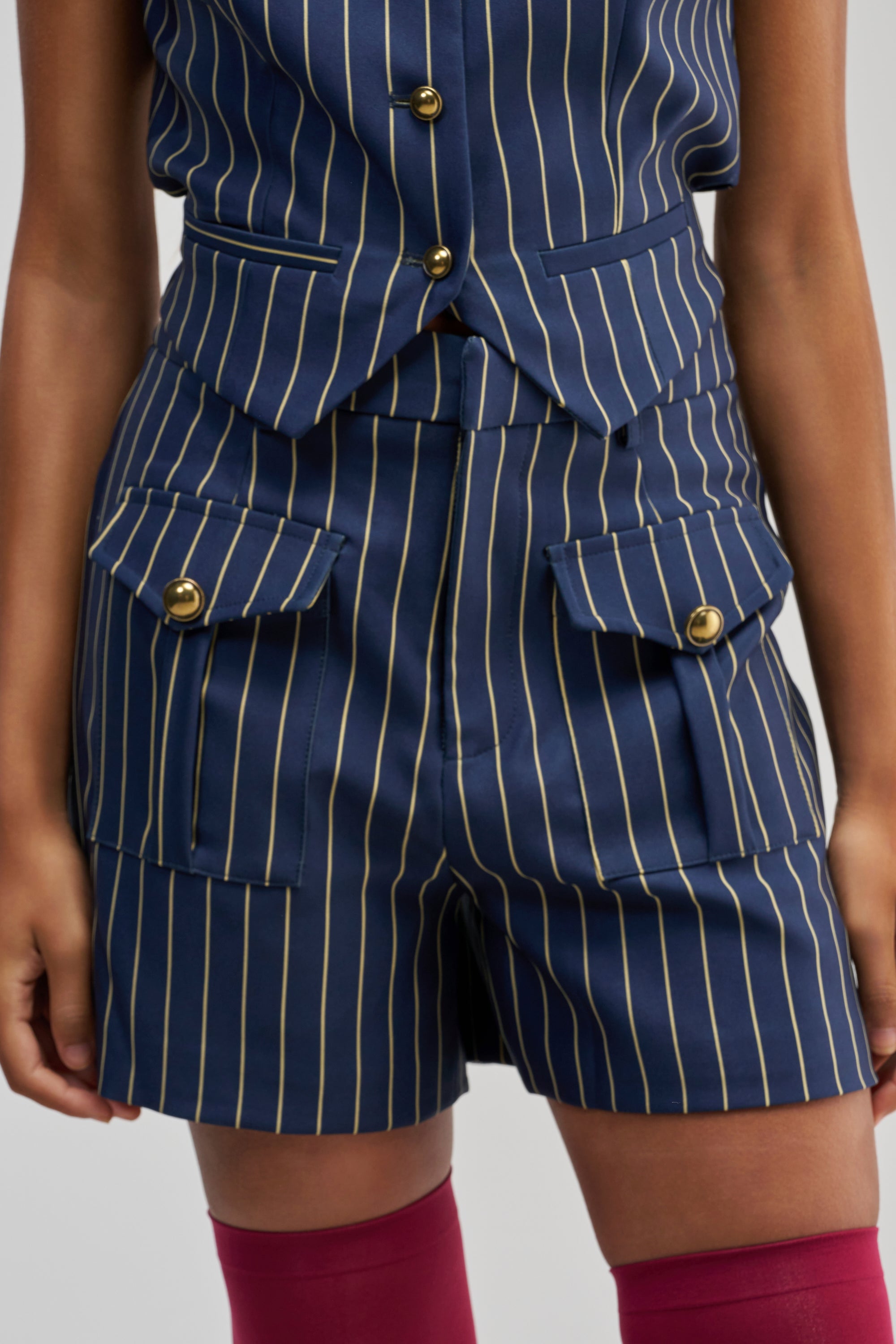 A person wearing the SUNDAY PINSTRIPE SHORTS and a matching navy blue pinstripe vest with gold buttons. Their hands are relaxed at their sides, and they are also wearing red thigh-high stockings.
