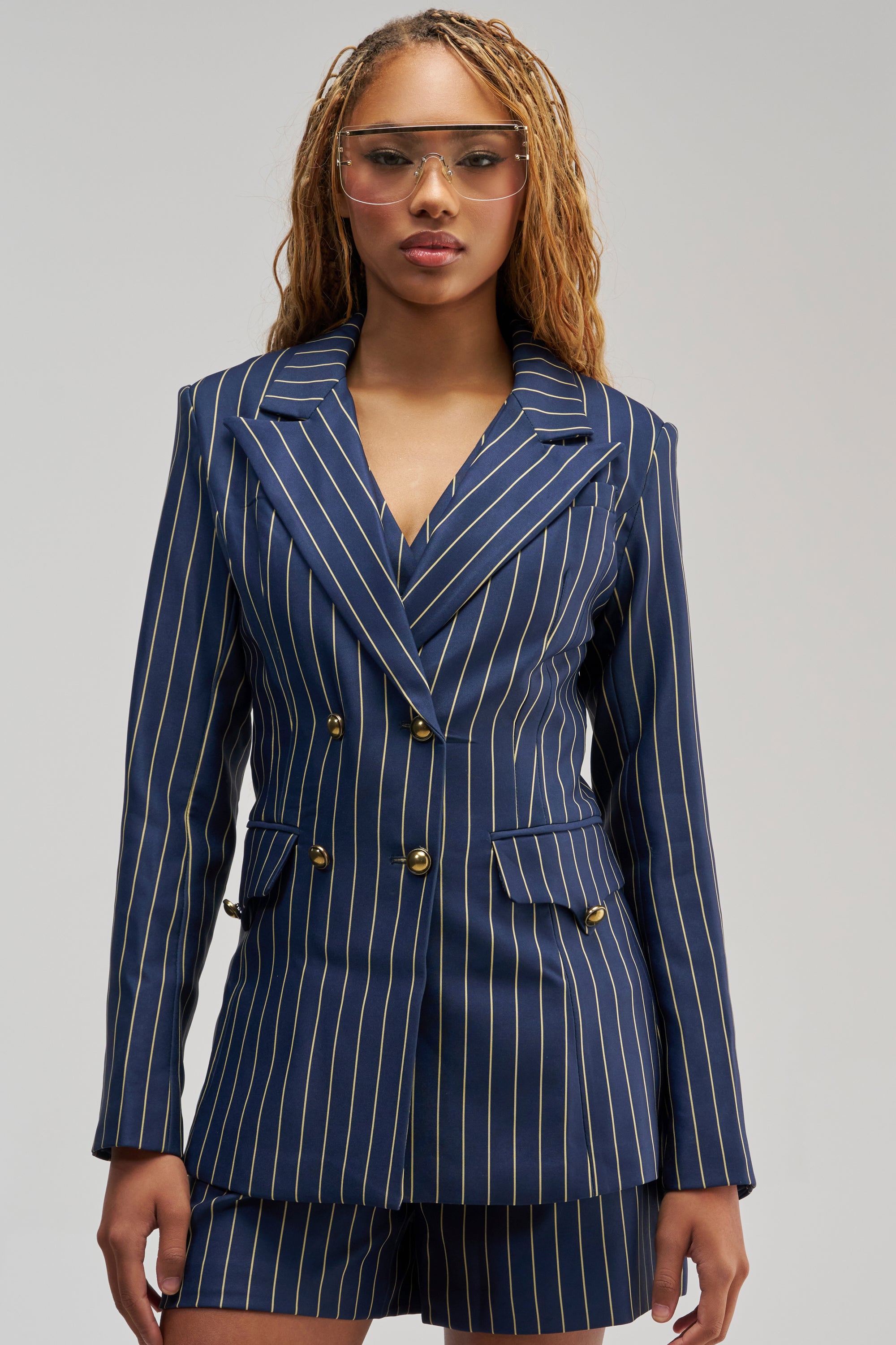 A woman stands facing forward against a plain light background, wearing the SUNDAY PINSTRIPE BLAZER—a navy blue double-breasted pinstripe blazer with gold buttons—paired with matching shorts. She has light brown braided hair and clear glasses.