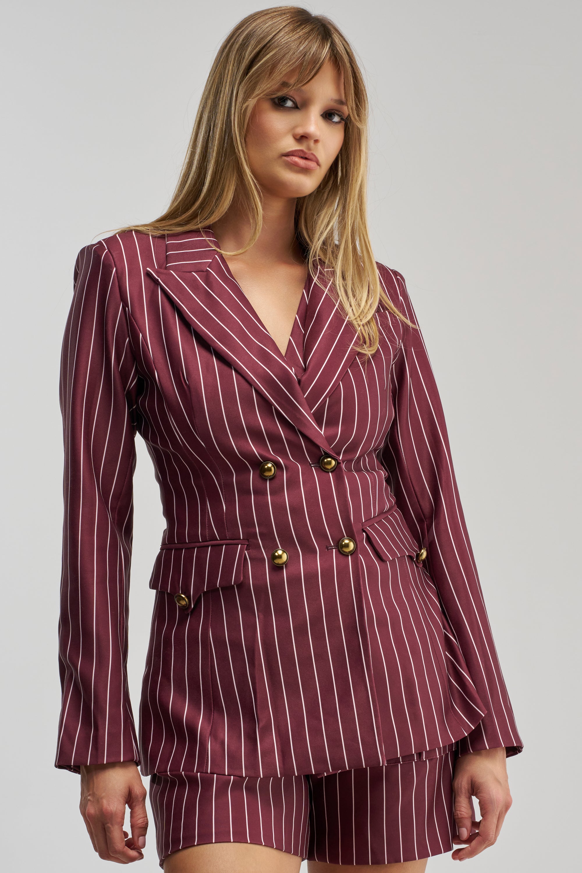 Wearing the SUNDAY PINSTRIPE BLAZER IN BURGUNDY with matching shorts, a woman stands against a plain light gray background.