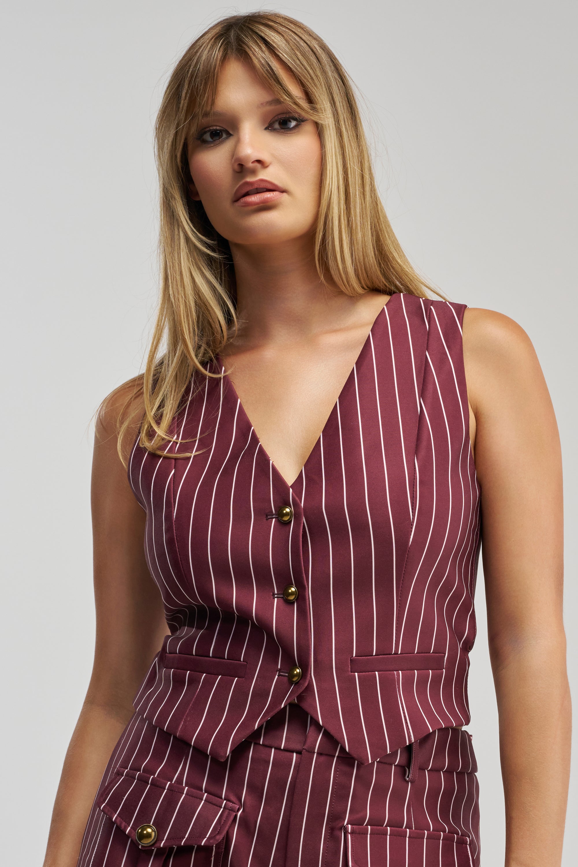 Wearing the SUNDAY PINSTRIPE VEST IN BURGUNDY, a woman with long blonde hair showcases this tailored, sleeveless piece with gold buttons and matching pants against a plain light background—an ideal look for modern office wear.