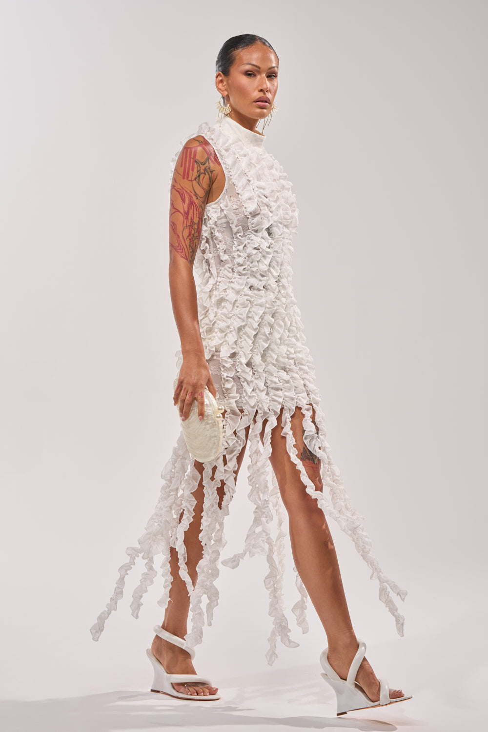 A woman with a visible arm tattoo wears the FOR THE FRILL OF IT MINI DRESS IN WHITE—a sleeveless, textured lace mini dress with long, ruffled hem strips. She holds a small white clutch and wears white heeled sandals against a plain backdrop.