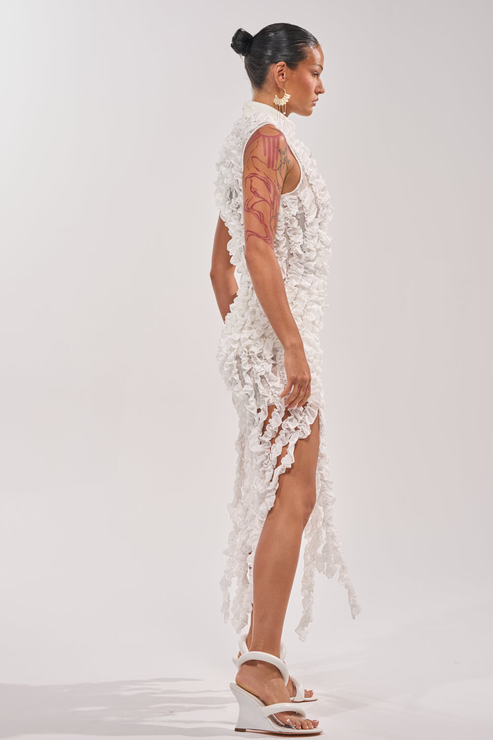 A person stands in profile wearing the FOR THE FRILL OF IT MINI DRESS IN WHITE featuring a mock neck, ruffled edges, and high slits, paired with white heels. Their hair is in a bun and they have a red abstract arm tattoo—ideal for a modern bridal party look.