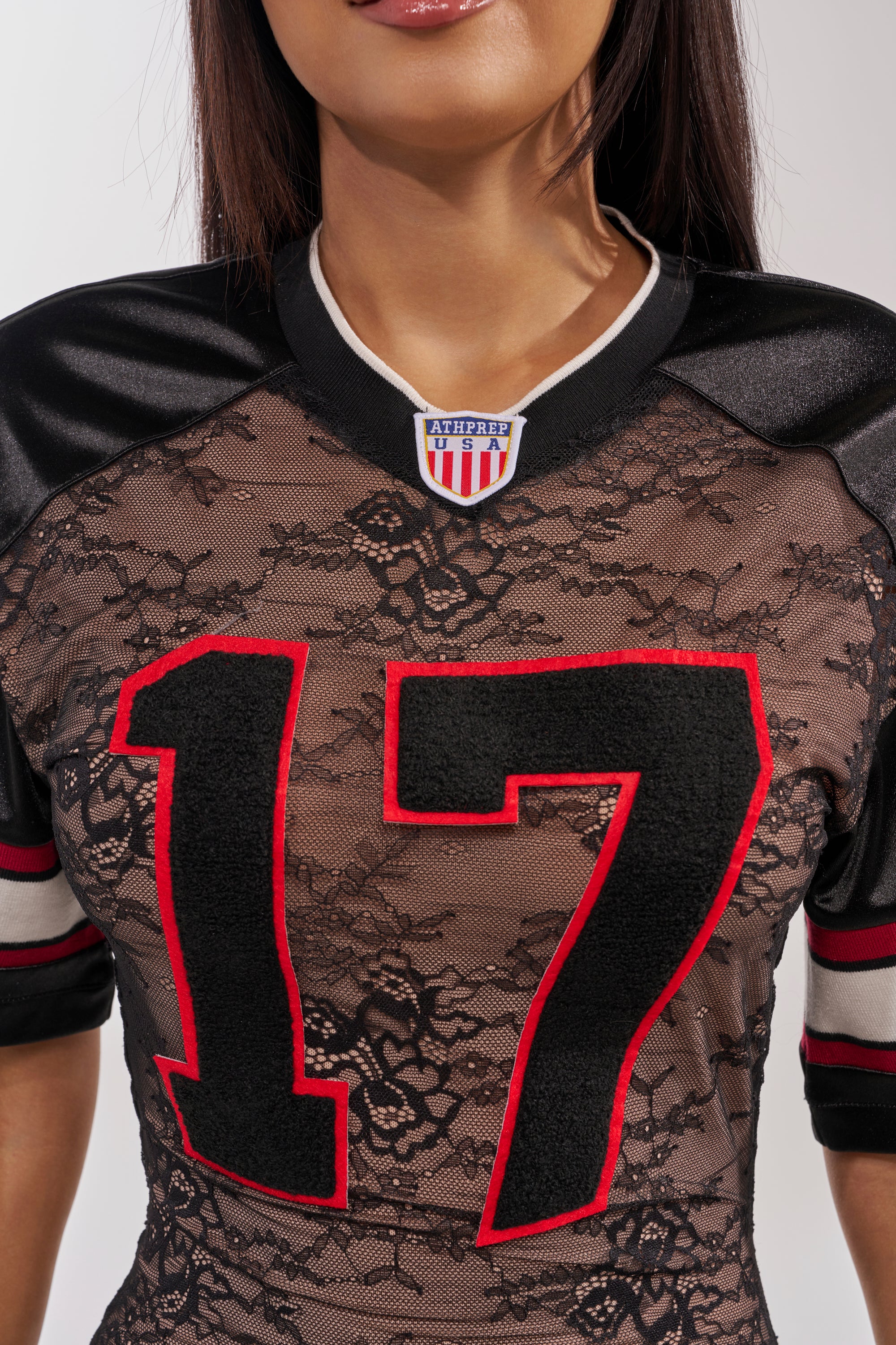A woman wears the LAYUP LACE JERSEY MIDI DRESS IN BLACK—a sporty black lace dress featuring red and black "17" numbers, shoulder stripes, and an "ATHLETE" patch near the collar. Her face is only partially visible. 5