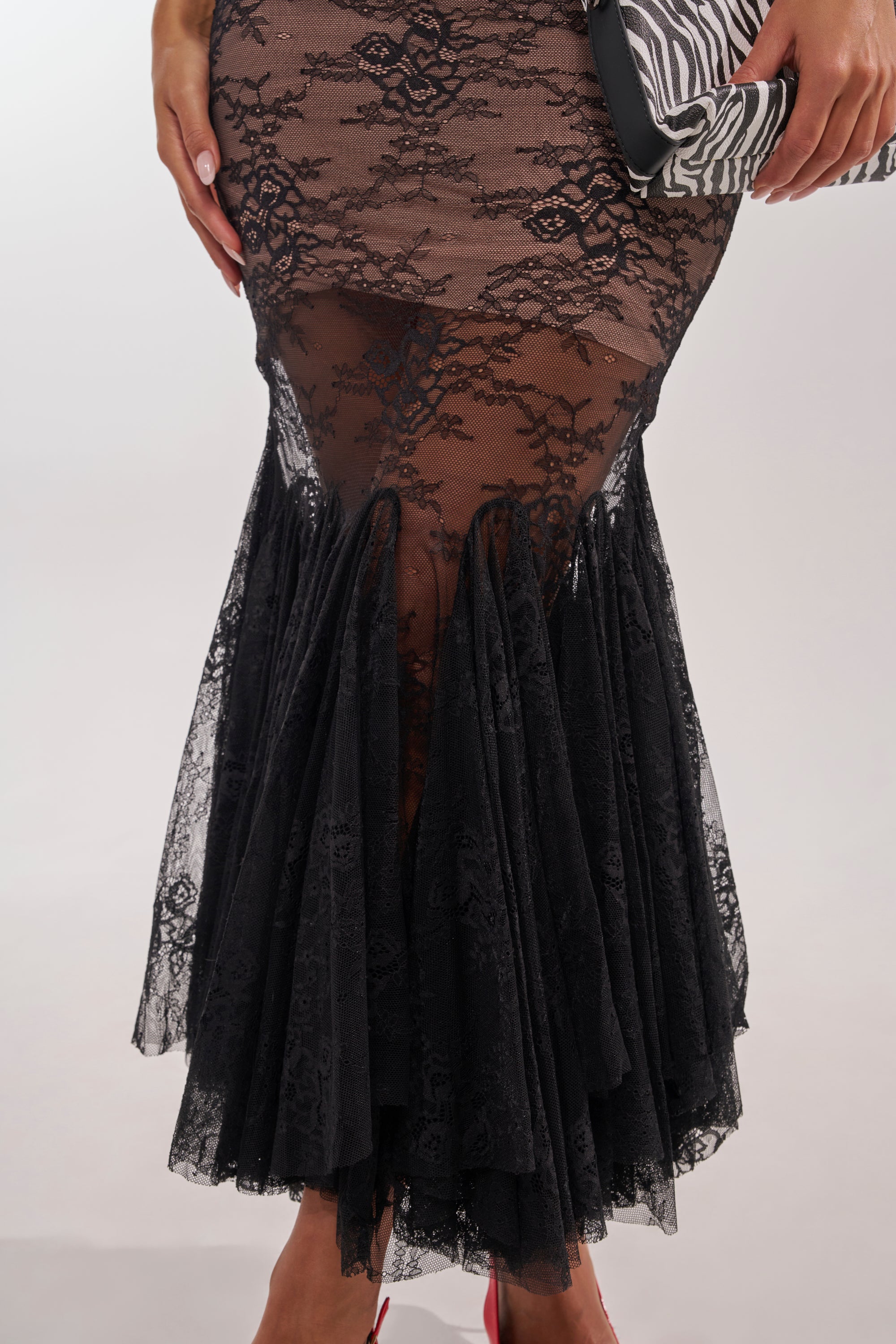The photo, cropped from the waist down, shows a person in the LAYUP LACE JERSEY MIDI DRESS IN BLACK—form-fitting and sheer with floral lace and ruffled knee details—holding a zebra-print clutch bag. 6
