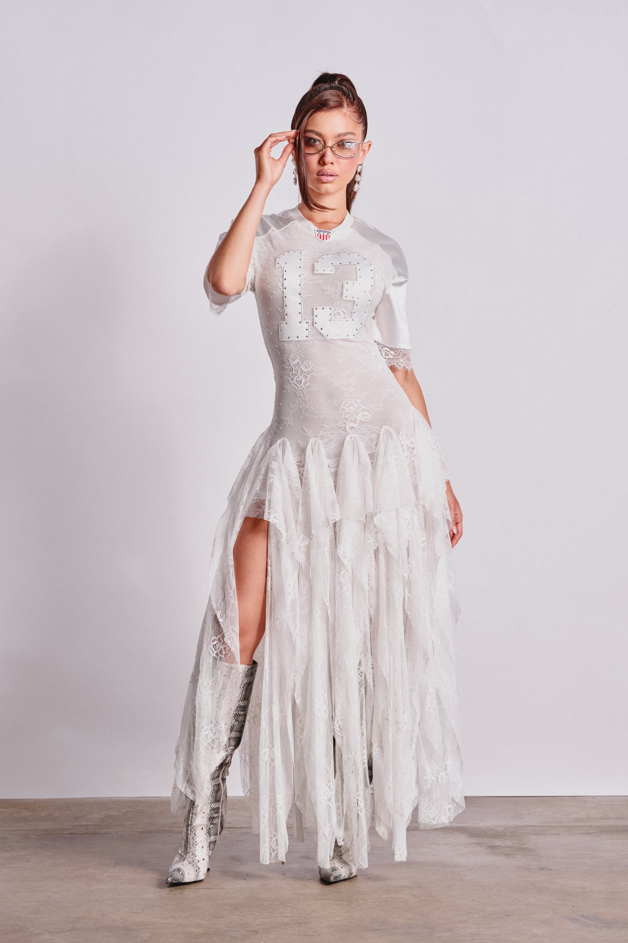 Wearing the KICK OFF LACE JERSEY MAXI DRESS IN WHITE with a high slit, clear glasses, and knee-high patterned boots, this confident pose against a plain background is perfect festival outfit inspiration. 0